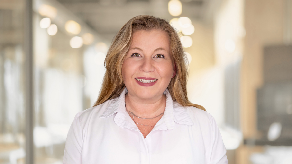 Smiling woman with long light brown hair wearing a white blouse, standing in a bright modern office environment.
