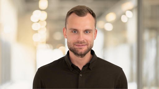 A man with short brown hair and a neatly trimmed beard standing in a modern office hallway, wearing a dark button-up shirt, looking directly at the camera with a calm and confident expression, photographed in landscape format with a softly blurred background and warm lights.