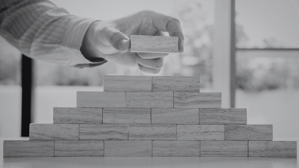 Hand placing a wooden block on top of a pyramid-shaped stack of wooden blocks on a table.