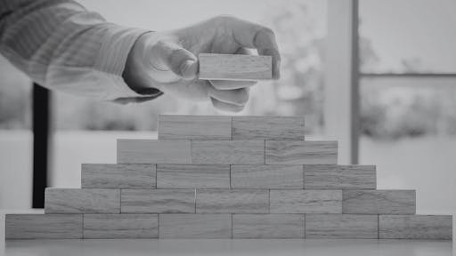 Hand placing a wooden block on top of a pyramid-shaped stack of wooden blocks on a table.