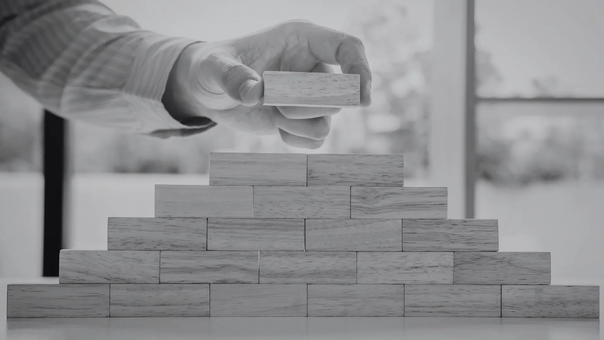 Hand placing a wooden block on top of a pyramid-shaped stack of wooden blocks on a table. Hand placing a wooden block on top of a pyramid-shaped stack of wooden blocks on a table.