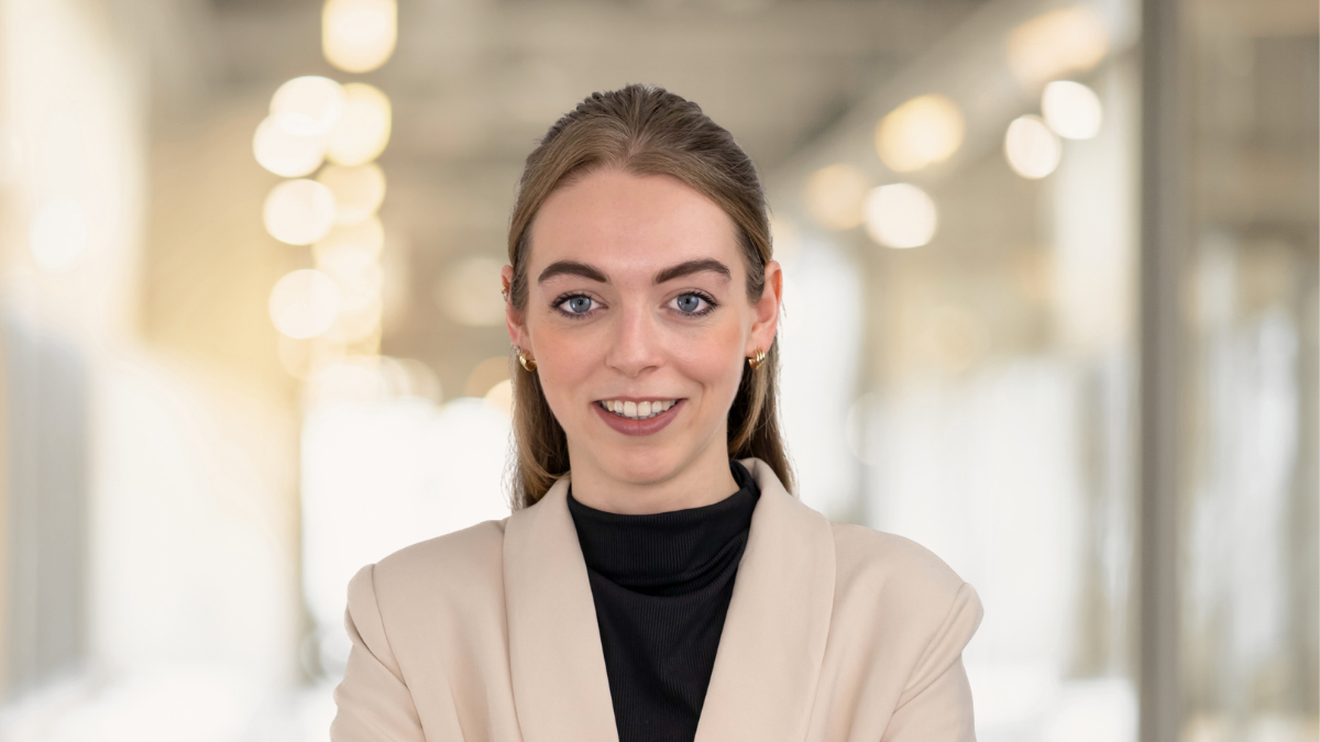 Smiling woman with long blonde hair wearing a beige blazer and a black shirt, standing in a bright modern office environment.