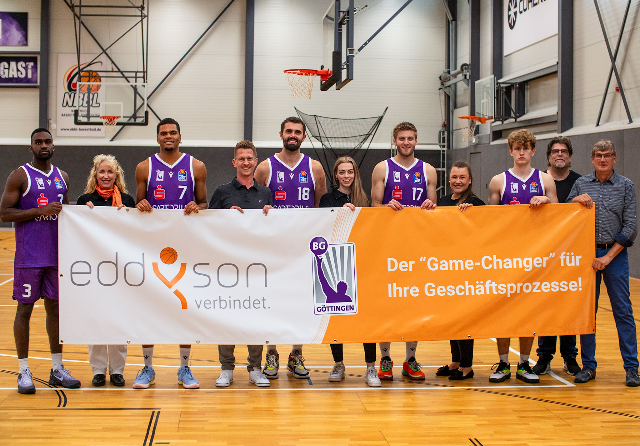 A group of basketball players and staff are standing in a line on a basketball court. They are holding a large banner with the logos "eddyson verbindet" and "BG Göttingen", along with some text in German. The setting appears to be a sports facility.
