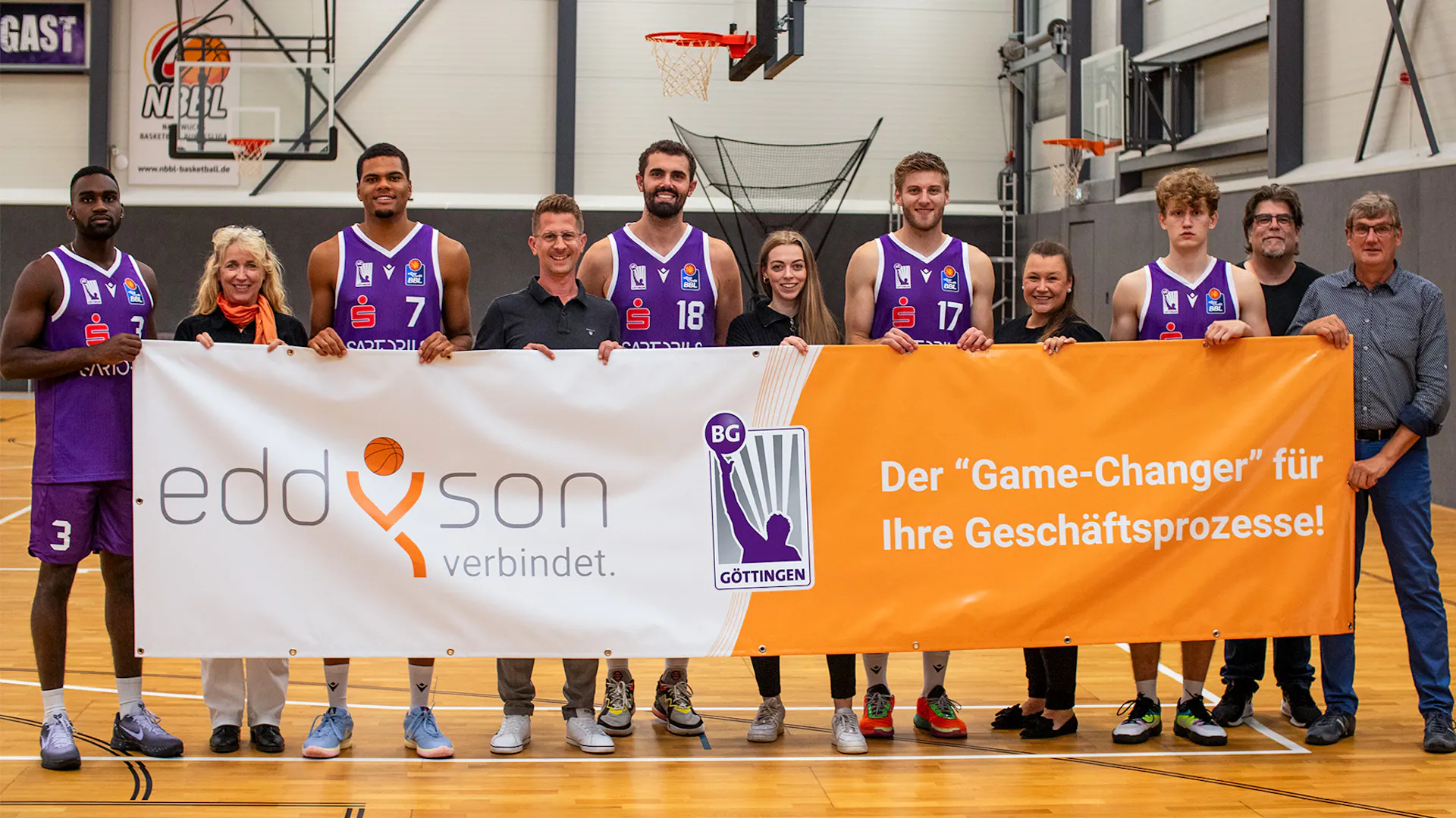A group of basketball players and staff are standing in a line on a basketball court. They are holding a large banner with the logos "eddyson verbindet" and "BG Göttingen", along with some text in German. The setting appears to be a sports facility. A group of basketball players and staff are standing in a line on a basketball court. They are holding a large banner with the logos "eddyson verbindet" and "BG Göttingen", along with some text in German. The setting appears to be a sports facility.