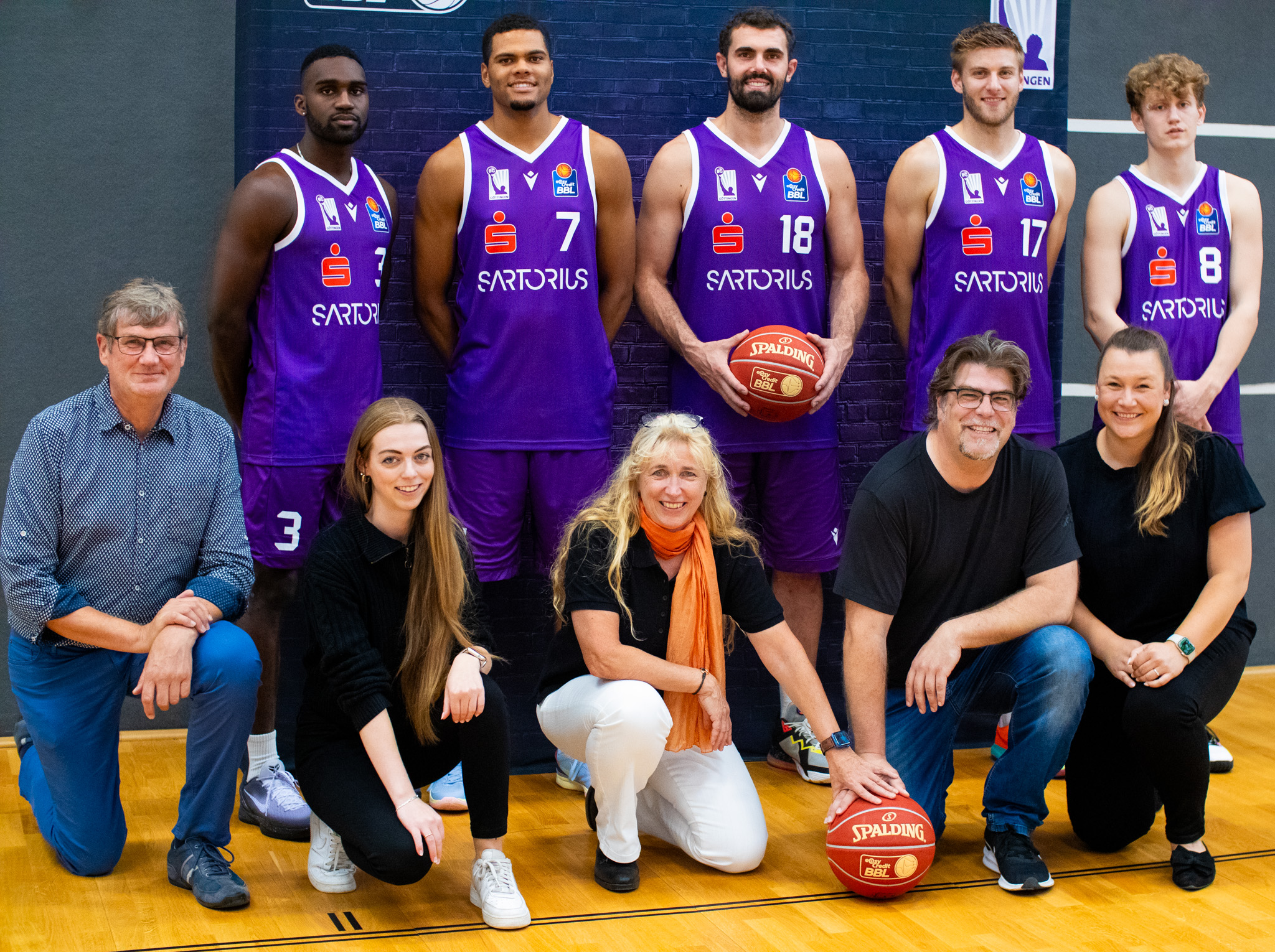 A basketball team and eddyson employees pose for a group photo on a basketball court. Four BG Göttingen basketball players stand in the back row wearing purple jerseys and holding a basketball. Five eddyson employees are kneeling in front of them. They are smiling into the camera. Behind them is a banner with a dark background.