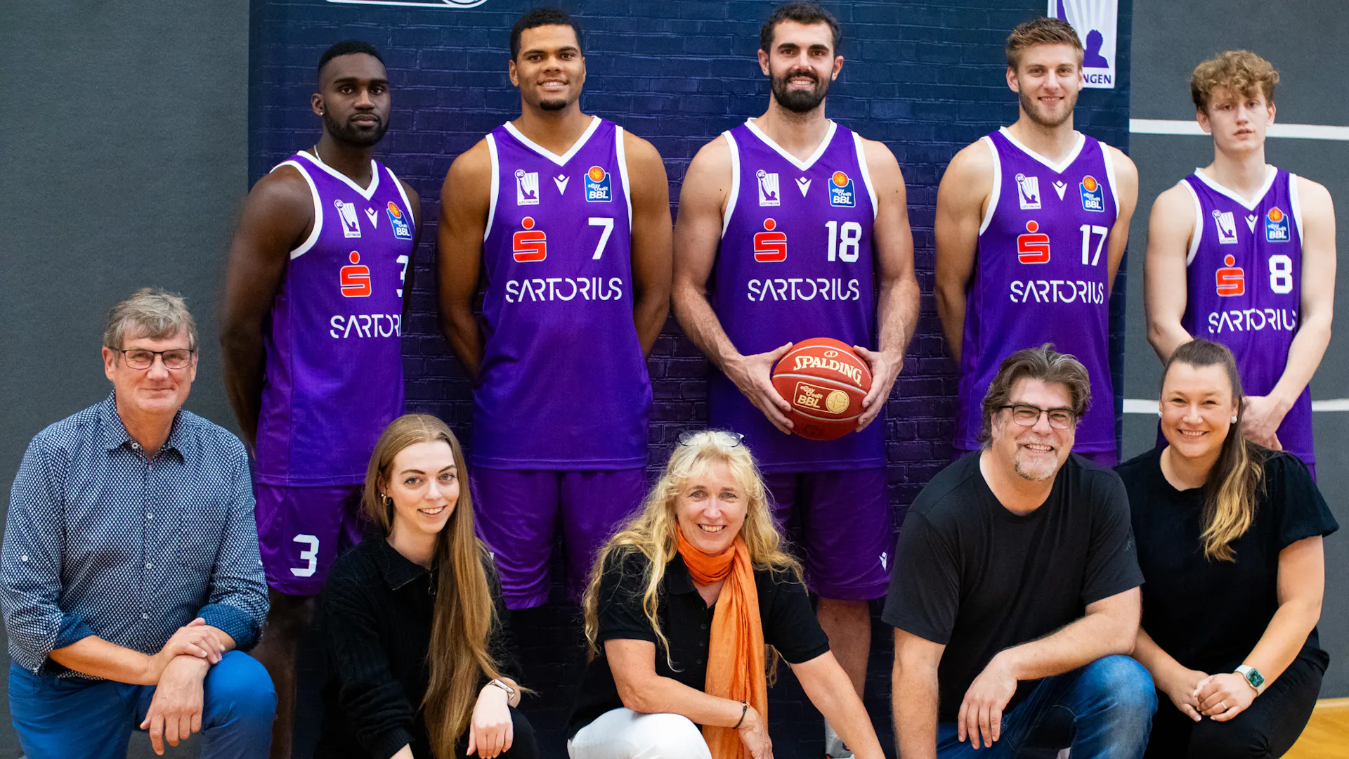A basketball team and eddyson employees pose for a group photo on a basketball court. Four BG Göttingen basketball players stand in the back row wearing purple jerseys and holding a basketball. Five eddyson employees are kneeling in front of them. They are smiling into the camera. Behind them is a banner with a dark background. A basketball team and eddyson employees pose for a group photo on a basketball court. Four BG Göttingen basketball players stand in the back row wearing purple jerseys and holding a basketball. Five eddyson employees are kneeling in front of them. They are smiling into the camera. Behind them is a banner with a dark background.