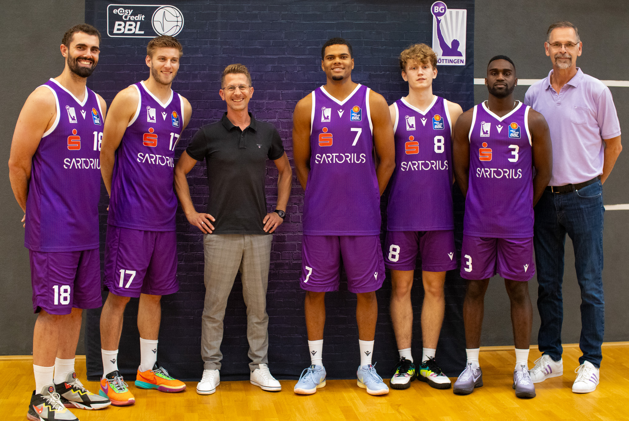 A group of six basketball players in purple uniforms stand together with two men in casual attire. They pose in front of a brick wall with signage for "BG Göttingen" and "easyCredit BBL." The players, each a standout basketball player, have their respective numbers visible on their uniforms.