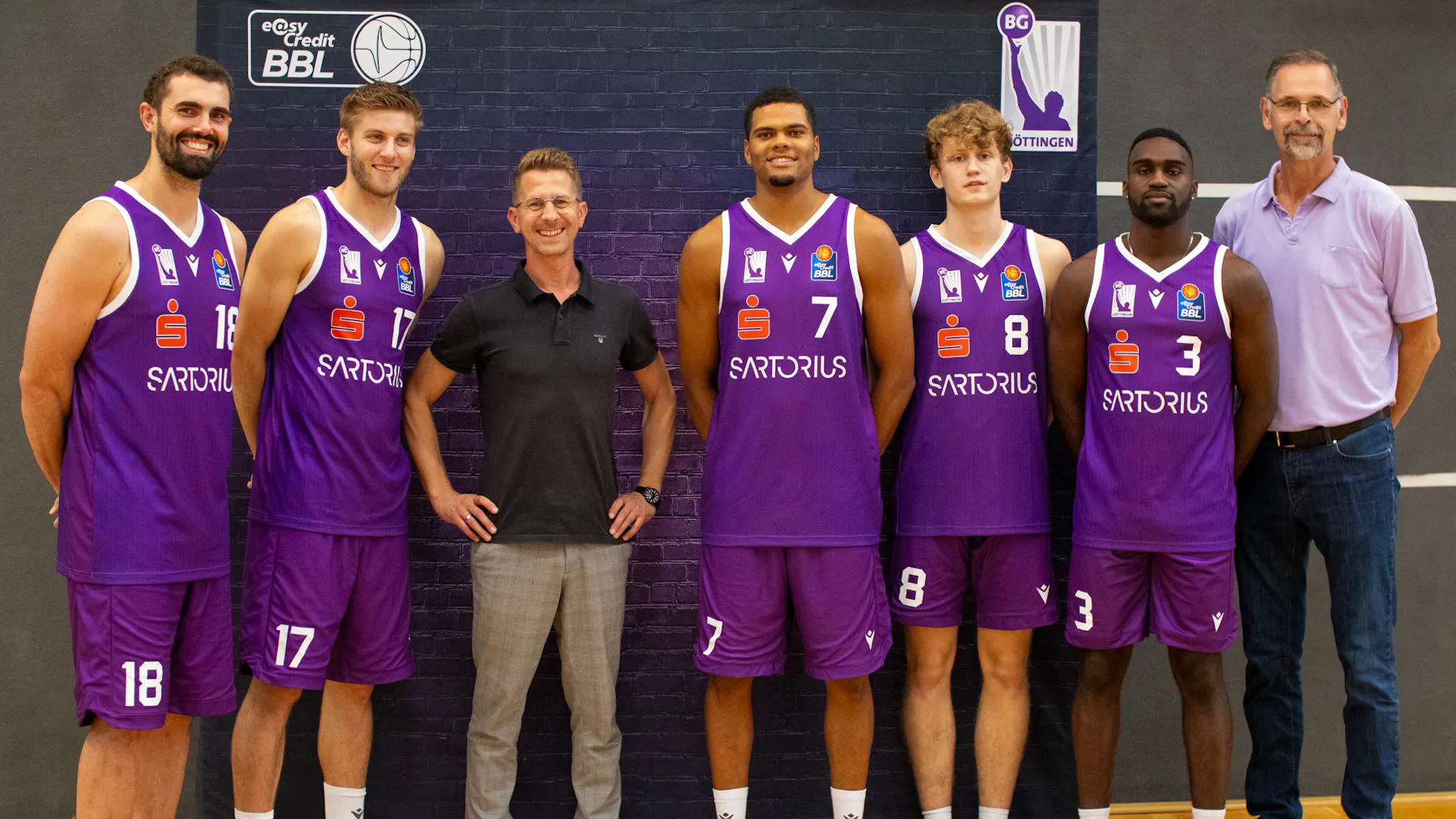 A group of six basketball players in purple uniforms stand together with two men in casual attire. They pose in front of a brick wall with signage for "BG Göttingen" and "easyCredit BBL." The players, each a standout basketball player, have their respective numbers visible on their uniforms. A group of six basketball players in purple uniforms stand together with two men in casual attire. They pose in front of a brick wall with signage for "BG Göttingen" and "easyCredit BBL." The players, each a standout basketball player, have their respective numbers visible on their uniforms.