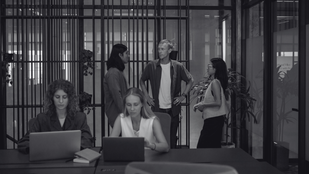 Black-and-white photo of a modern office. Two people are working on laptops at a conference table in the foreground, while three colleagues are standing and talking in a glass-walled meeting area in the background. The space features contemporary architecture with glass partitions, vertical metal frames, and indoor plants.