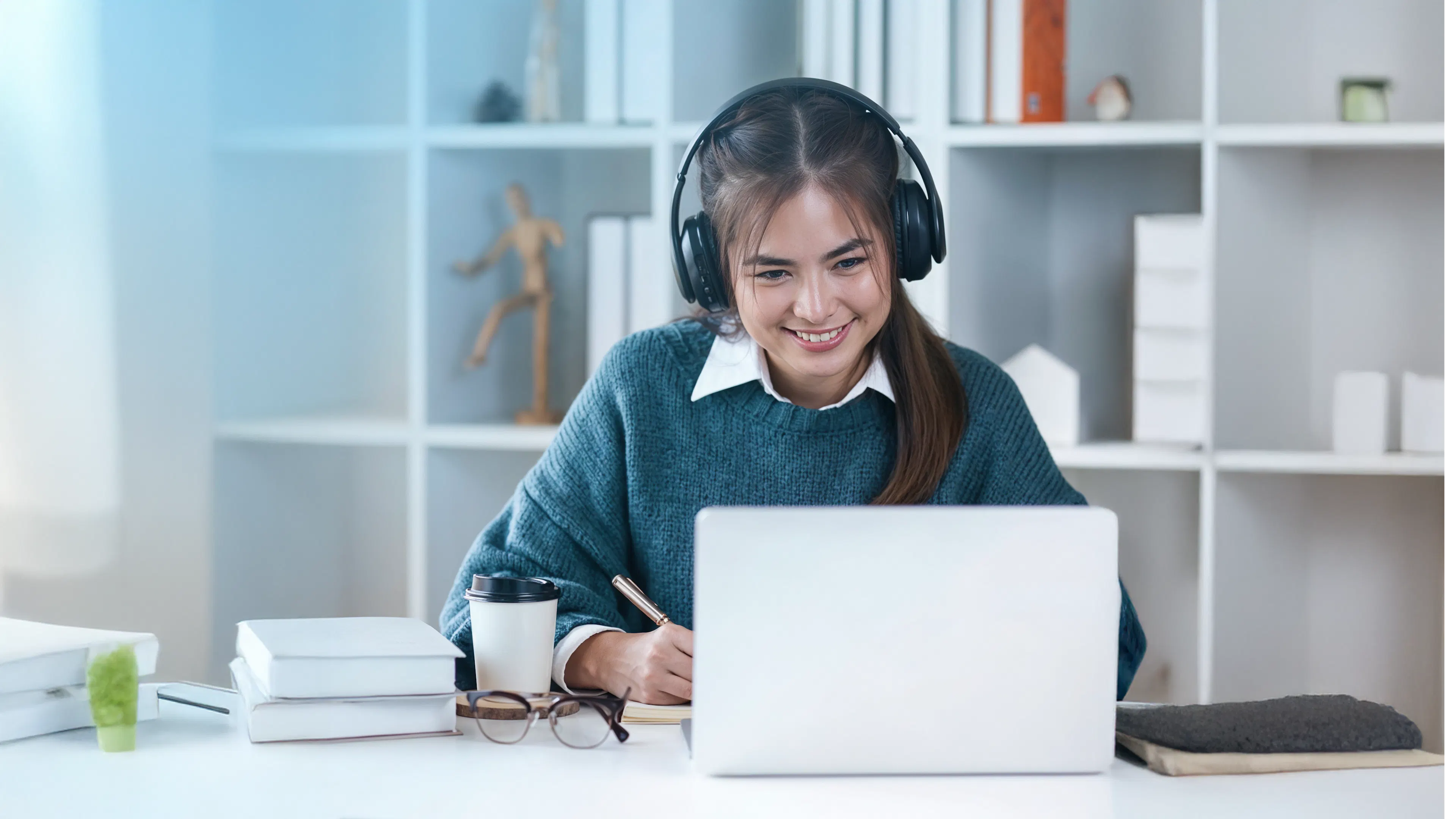 A young woman sits at a desk, smiling while wearing headphones and looking at a laptop. She takes notes, conveying focus and engagement. Books and a coffee cup are nearby.