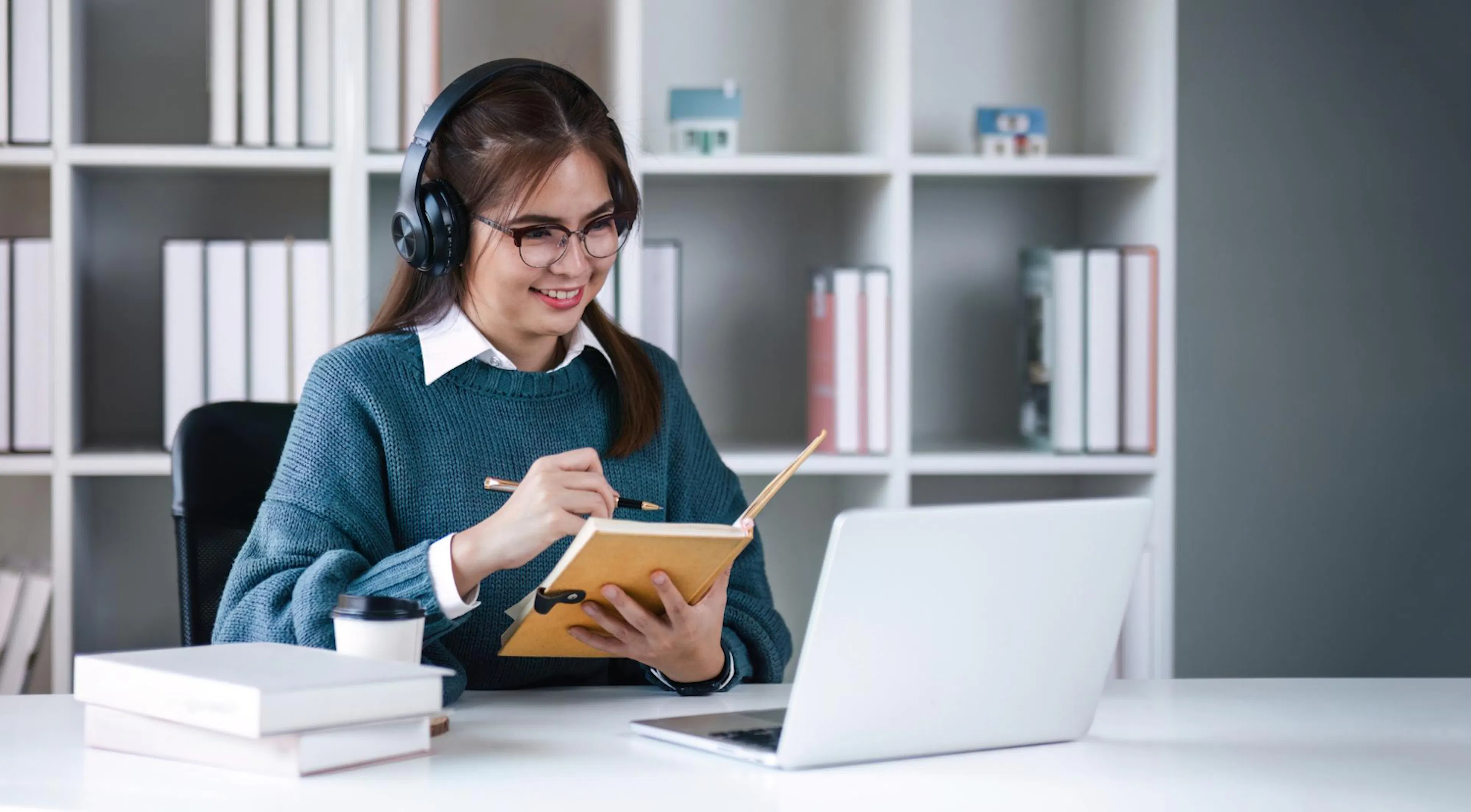 A woman sits at a desk, wearing headphones and glasses, smiling and writing in a notebook. A laptop, coffee cup, and books are on the desk, with bookshelves behind her.