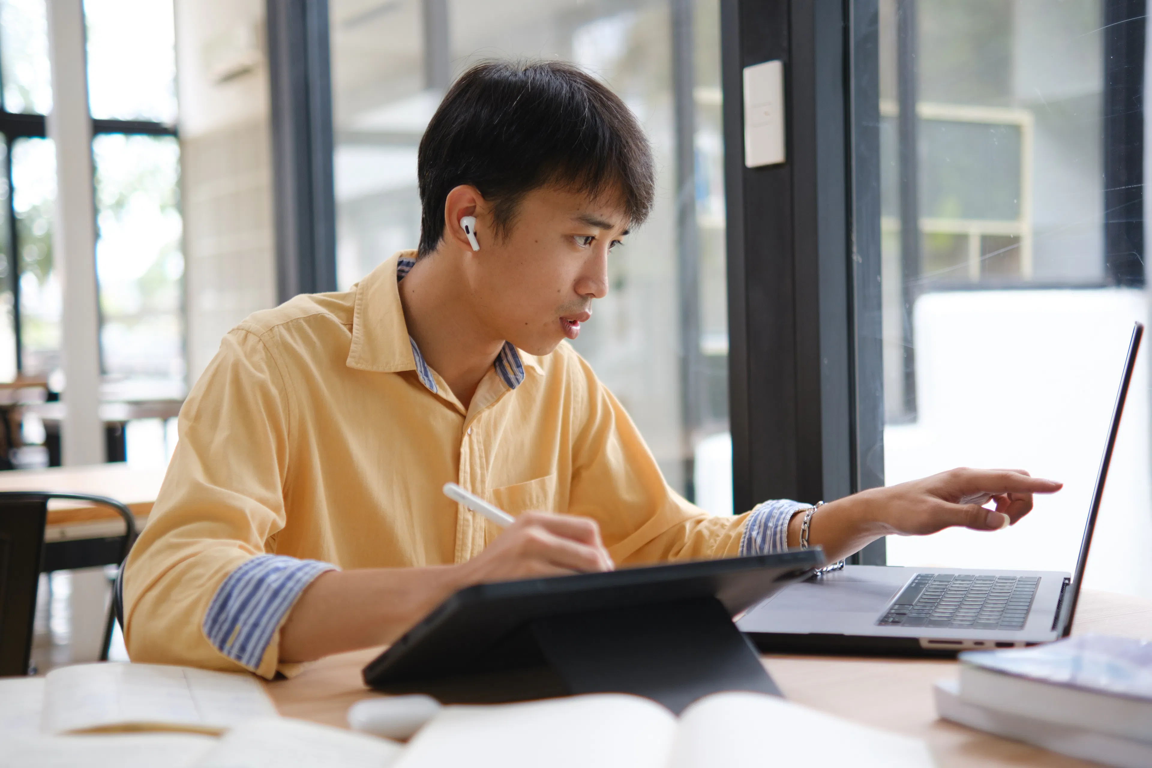Young man in a yellow shirt works intently on a laptop and tablet in a bright room, wearing earbuds, suggesting focus and productivity.