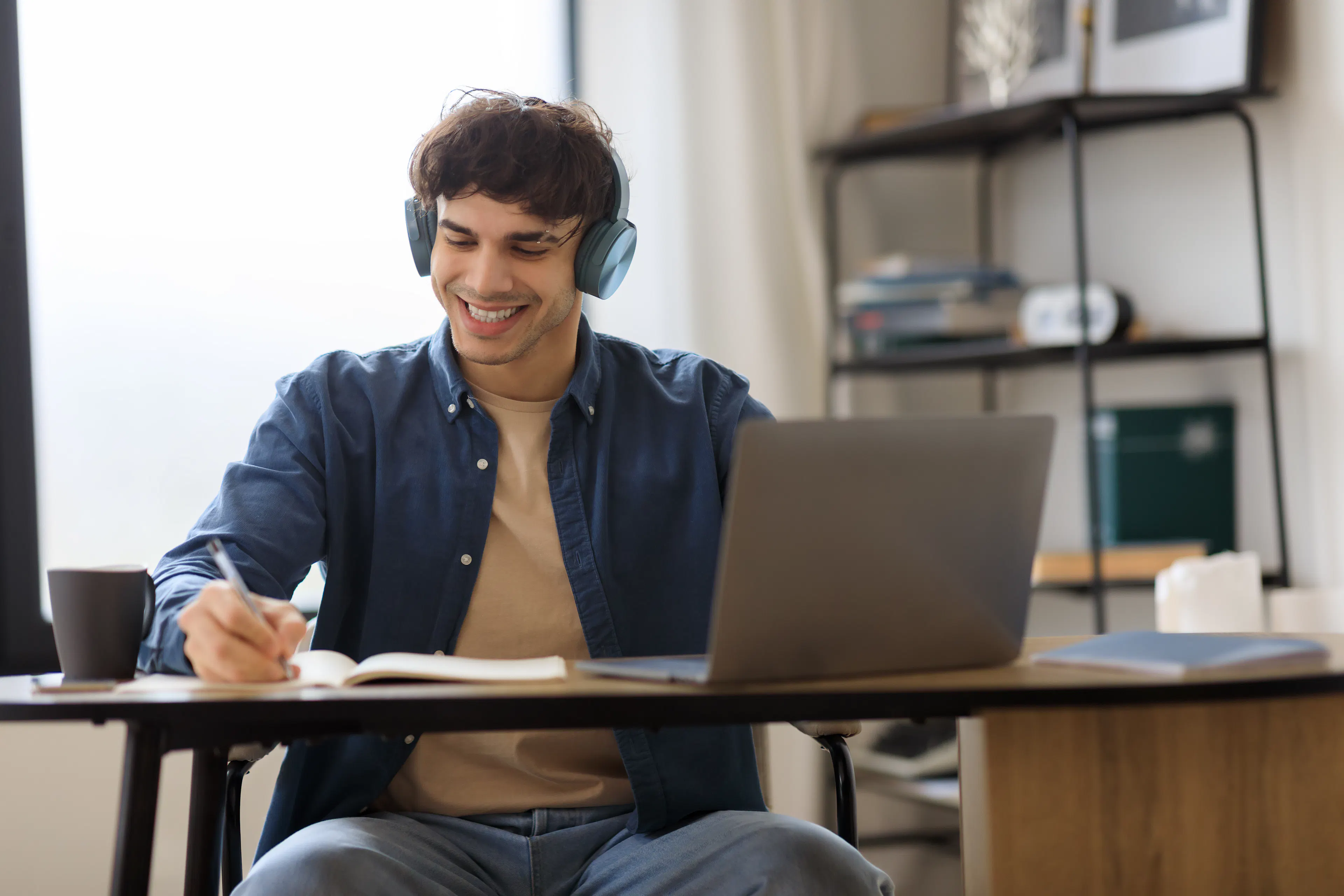 A young man wearing headphones smiles while writing in a notebook. He's seated at a desk with a laptop. The atmosphere is relaxed and focused.