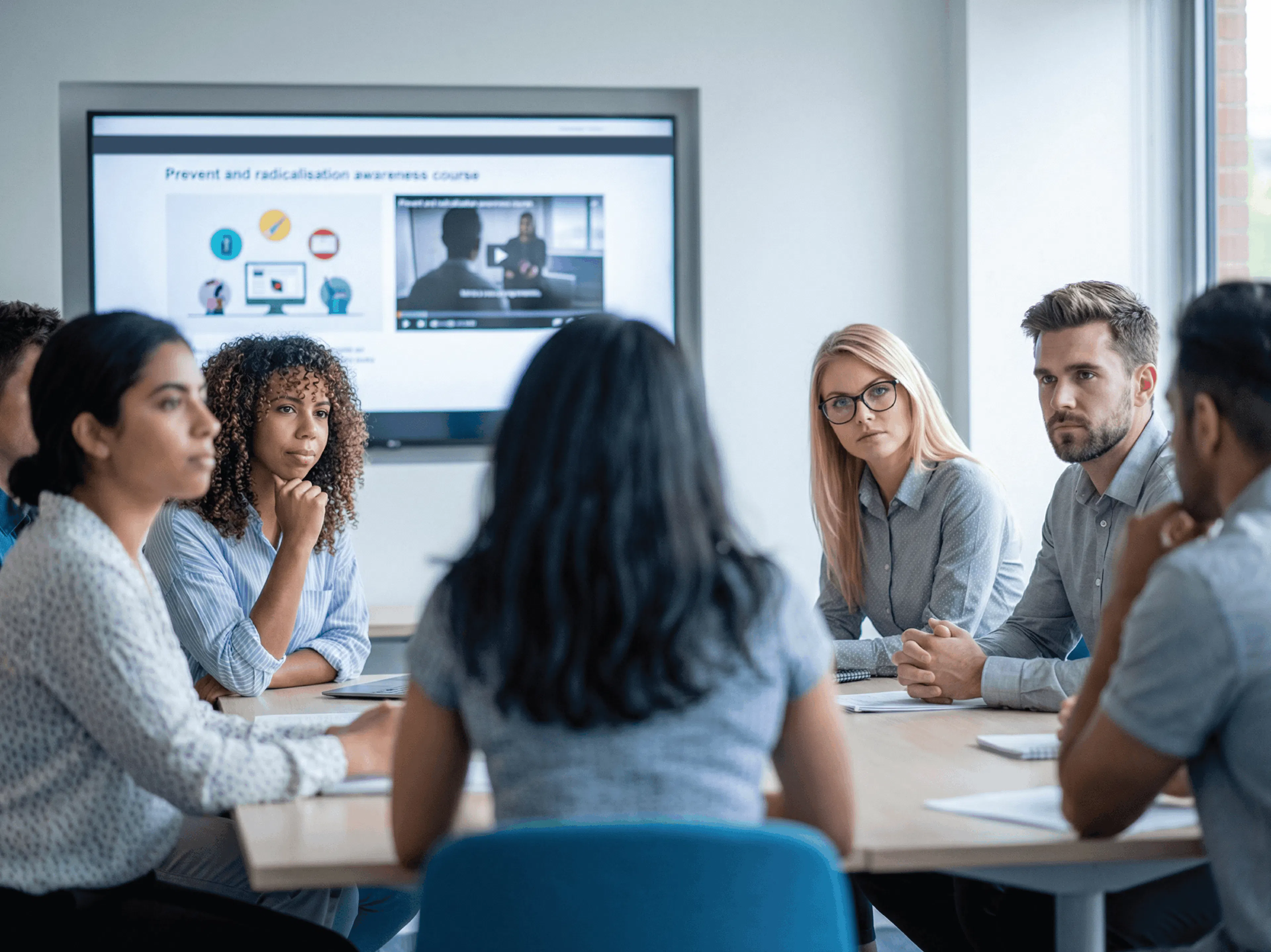 A diverse group of people sit attentively around a conference table, viewing a presentation titled "Prevent and Radicalisation Awareness Course" on a screen.