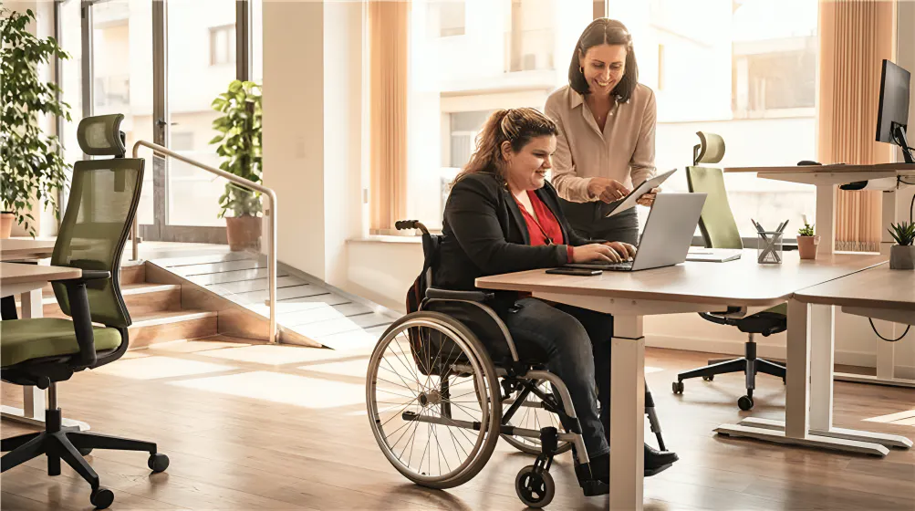 Two women are in a bright office, one in a wheelchair working on a laptop, the other standing beside her, smiling and pointing at the screen.