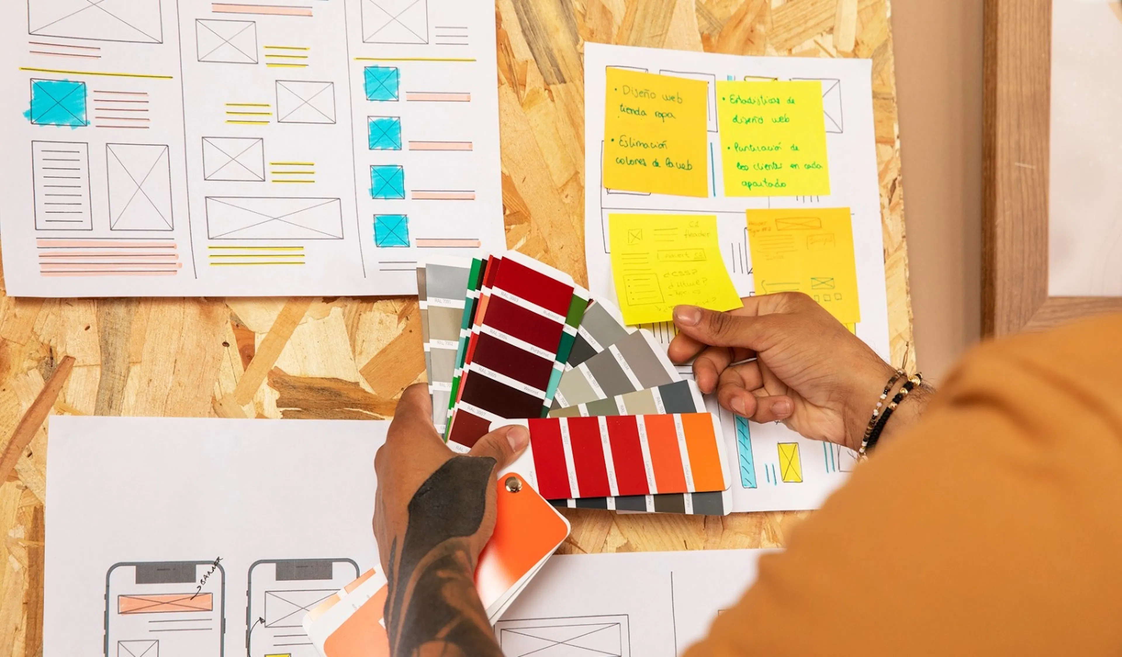 A person holds colour swatches against a bulletin board covered with wireframe sketches and sticky notes, conveying a creative and design-focused mood.