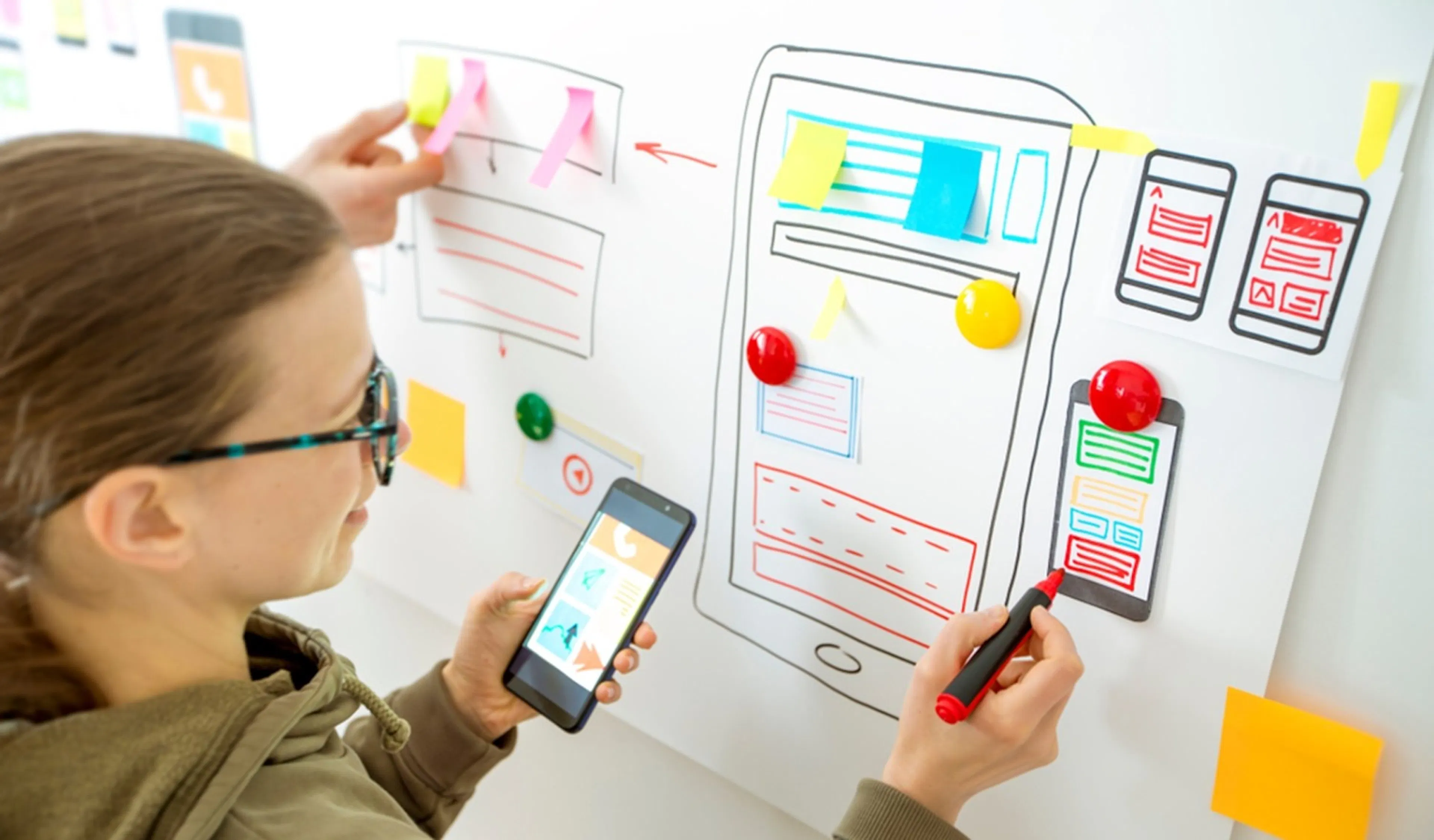 A person designs a mobile application interface on a whiteboard using colourful sticky notes and markers while holding a smartphone.