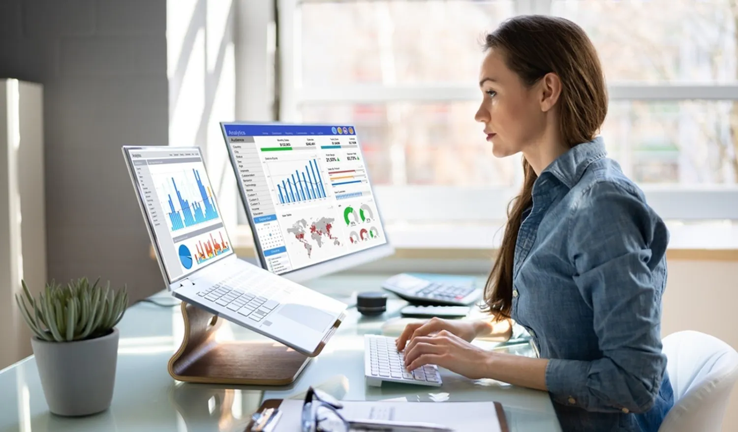 A woman in a denim shirt is focused on dual monitors displaying colourful data charts and graphs in an office. A small plant and some stationery are on her desk.