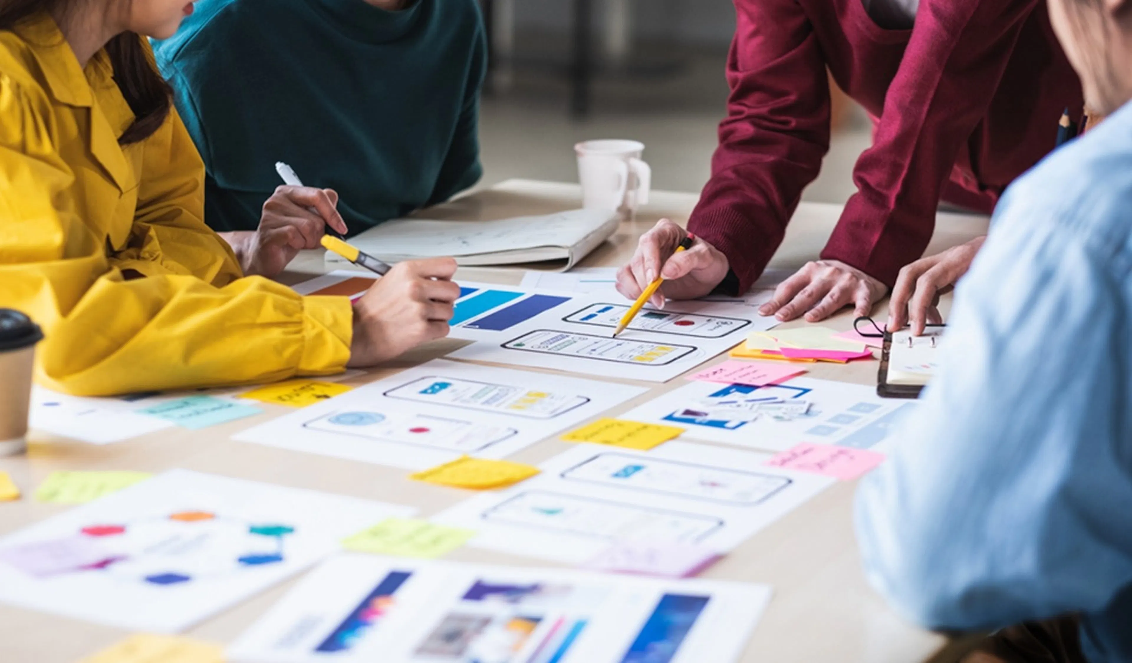 People collaborate around a table covered in design sketches and colourful sticky notes, suggesting teamwork and creativity in a lively office setting.