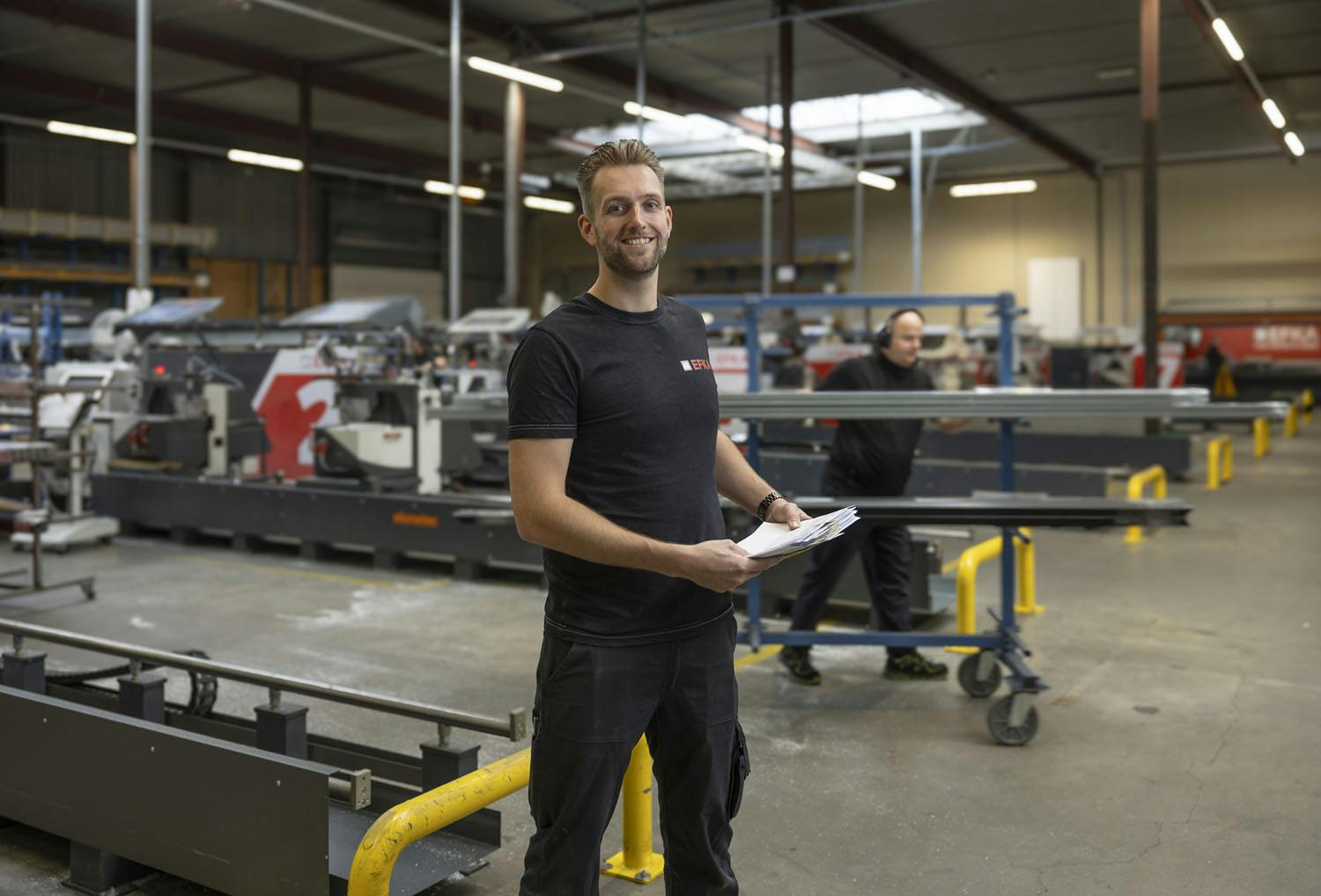 Medewerker in industriële hal, lachend naar de camera met papieren in hand