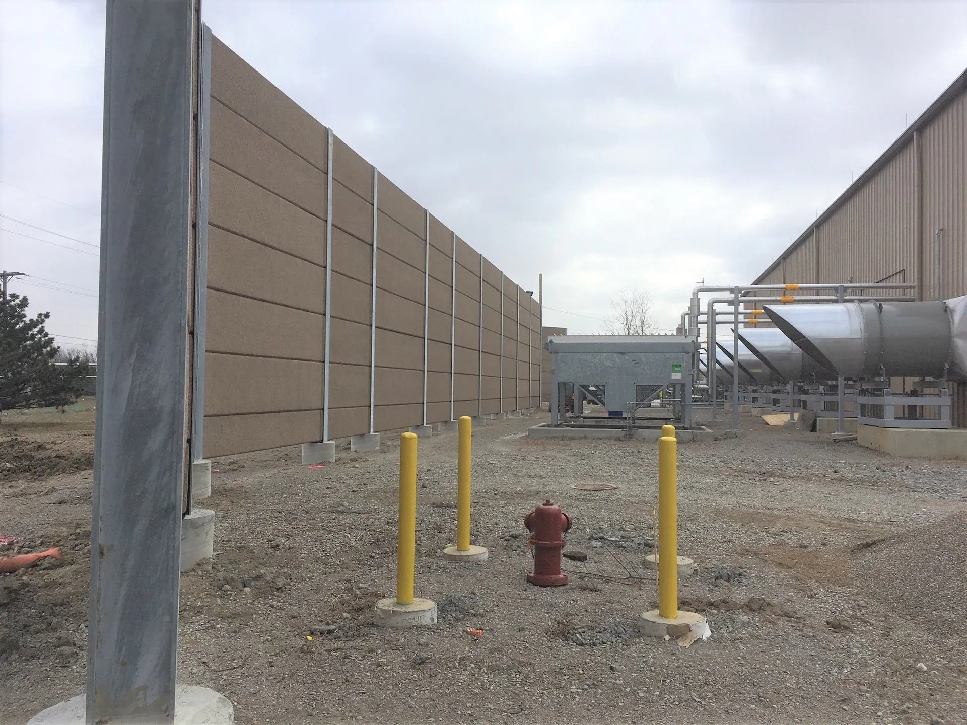 Tan noise wall enclosing the renewable natural gas processing equipment, with bollards and a hydrant in the foreground