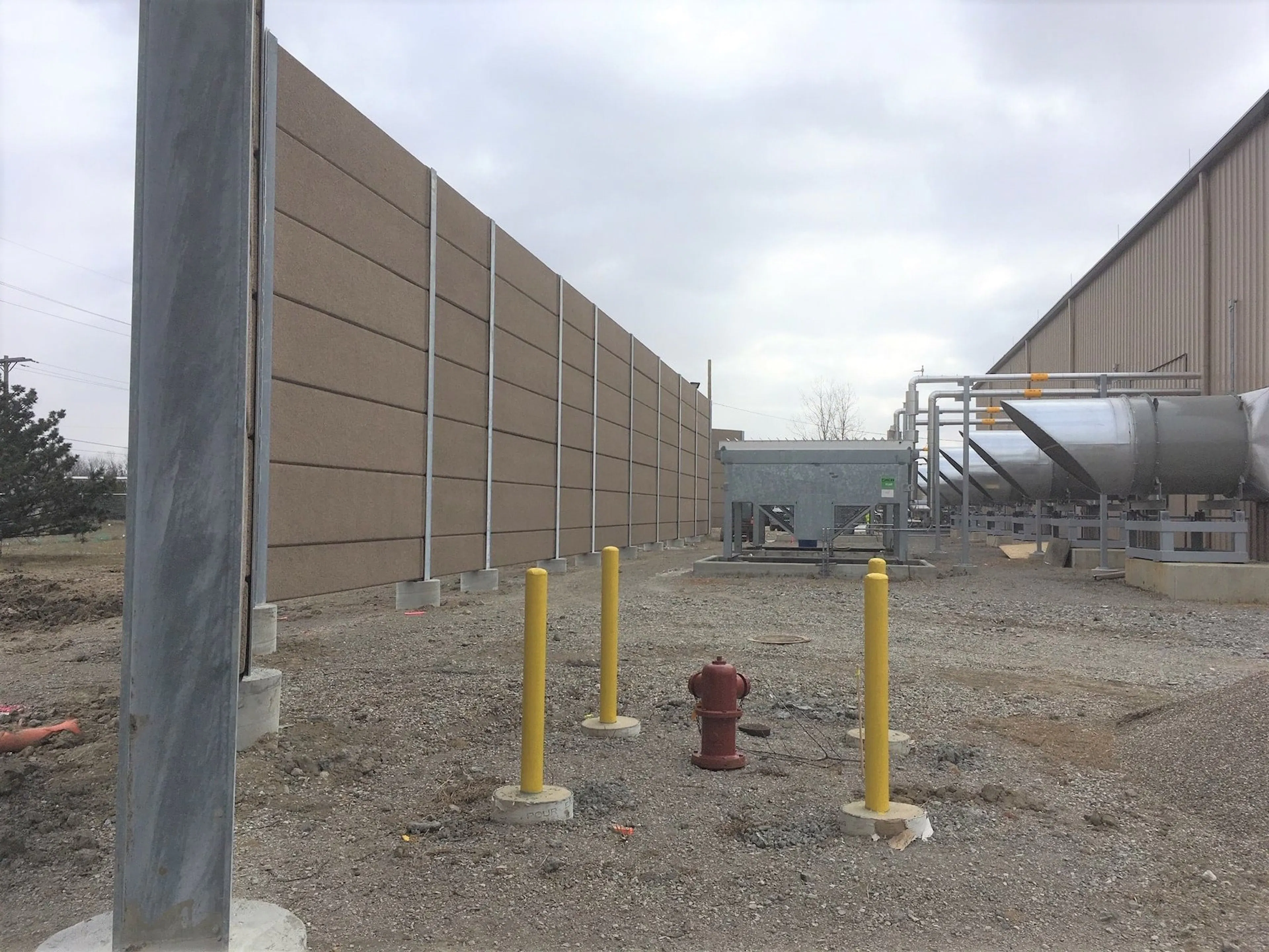 Tan noise wall enclosing the renewable natural gas processing equipment, with bollards and a hydrant in the foreground