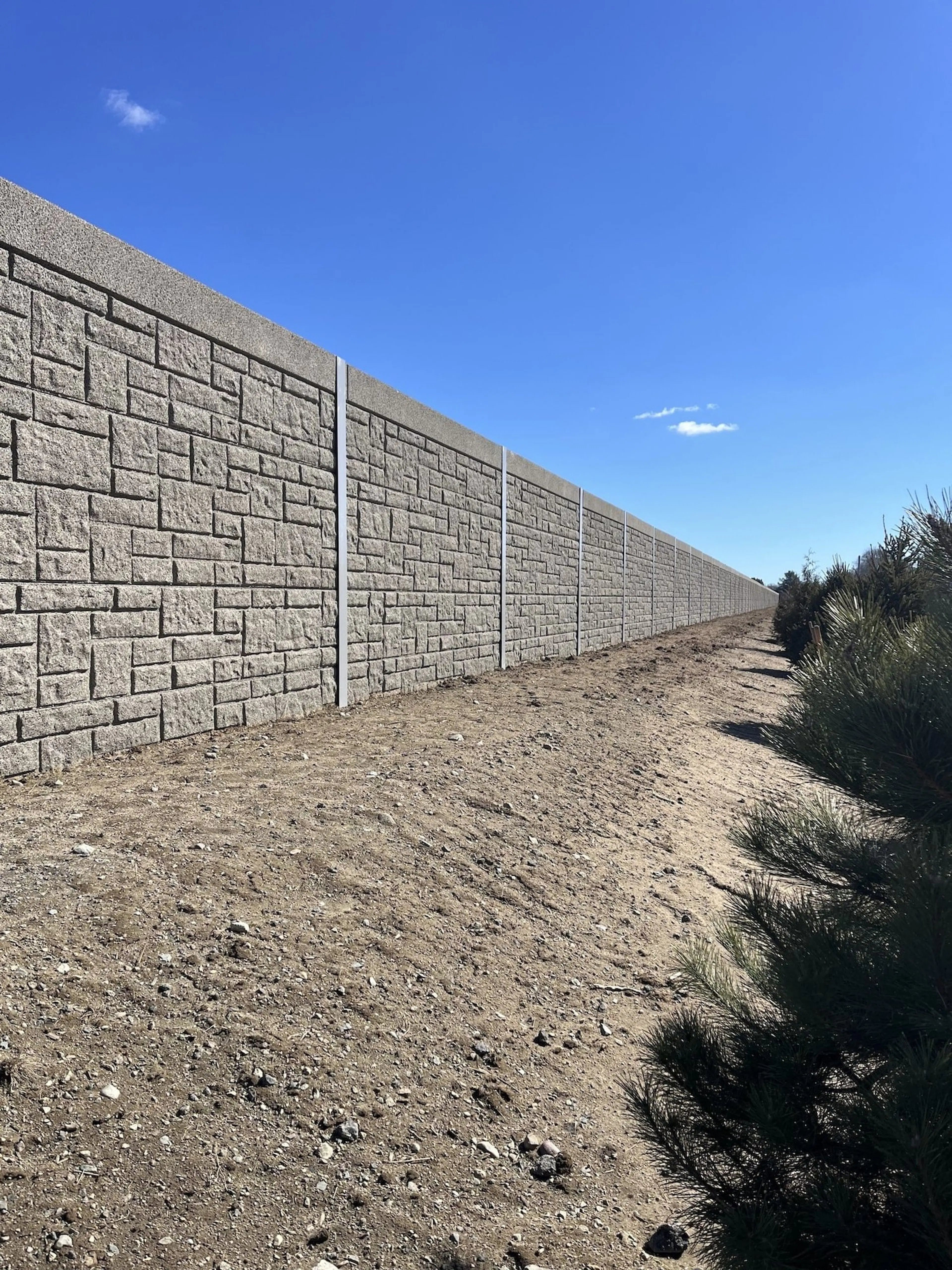Stone-textured sound wall running along a bare earth easement beside scrub at the airport boundary