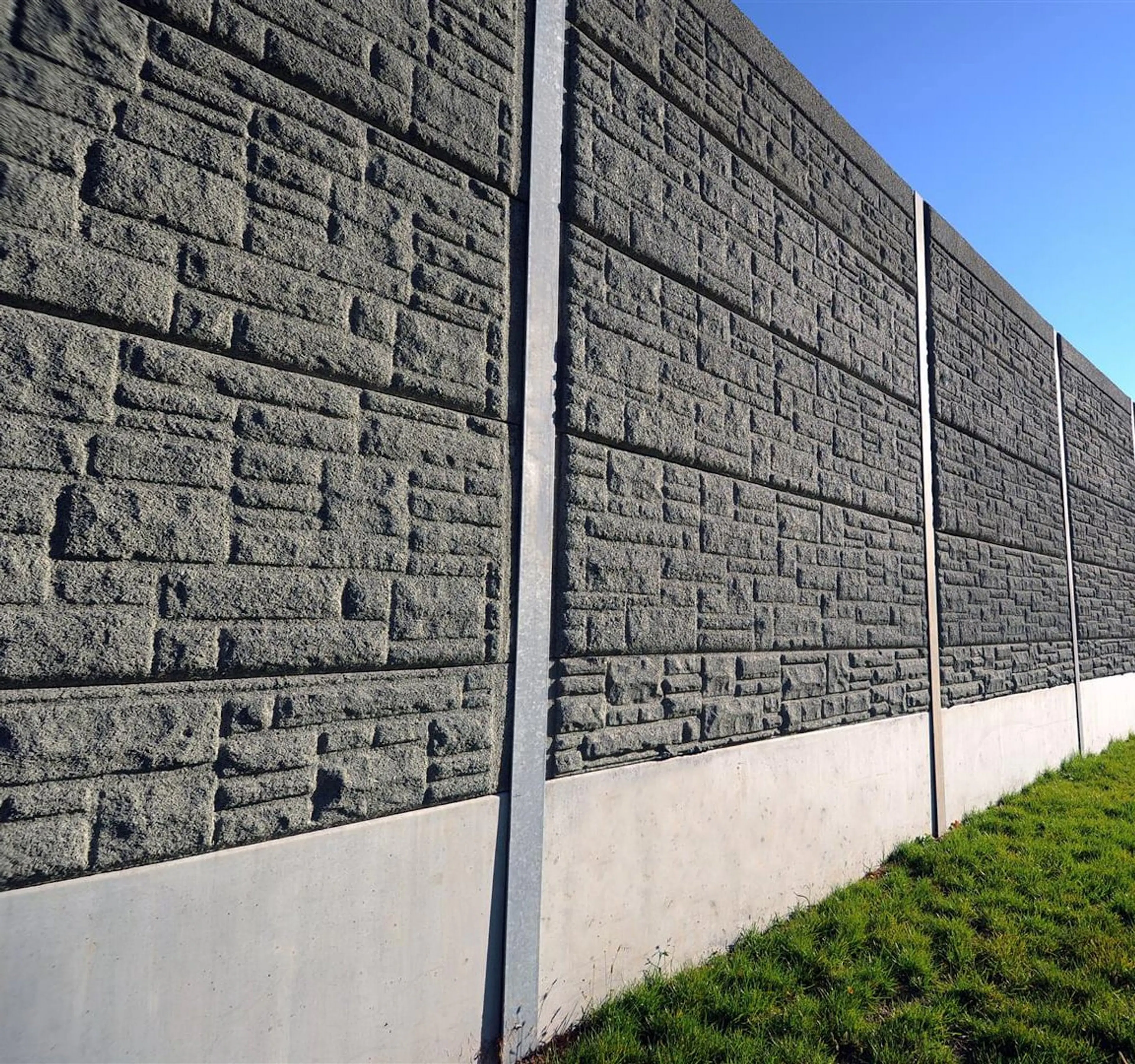 Grey stone-patterned absorptive noise barrier running beside a grassed shoulder and street lighting