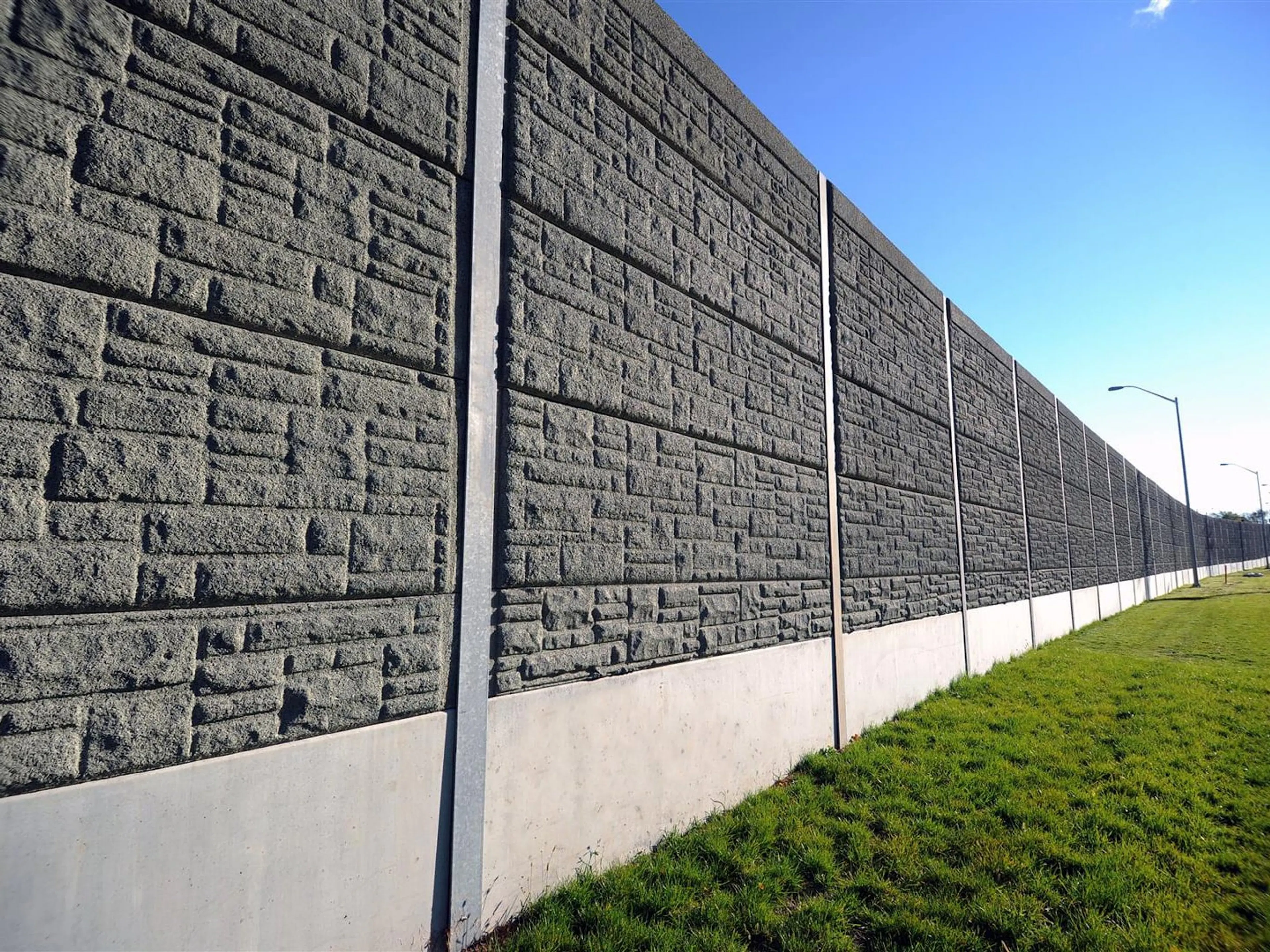 Grey stone-patterned absorptive noise barrier running beside a grassed shoulder and street lighting