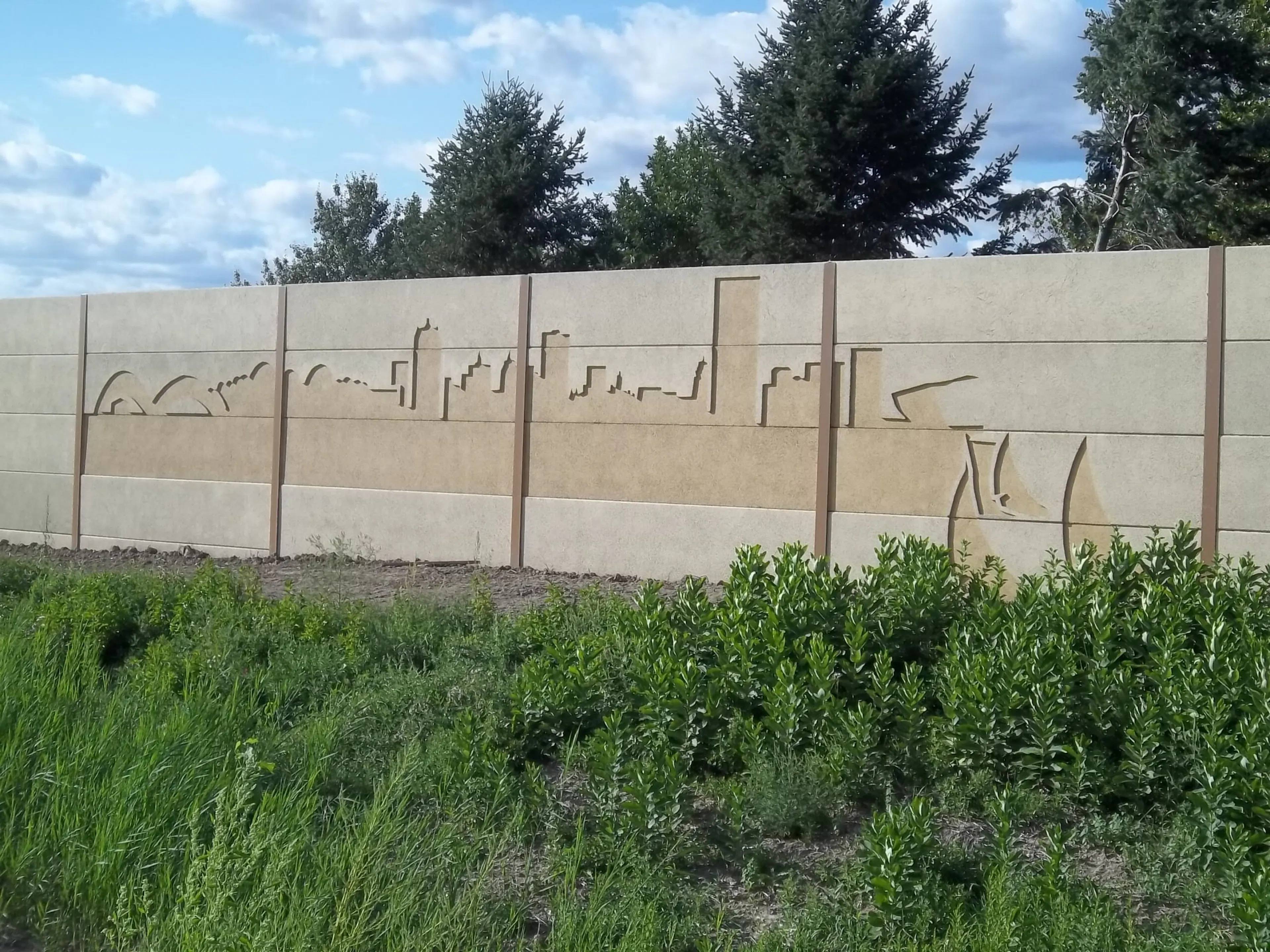 Milwaukee skyline mural recessed into the face of the noise wall above a bed of young shrubs