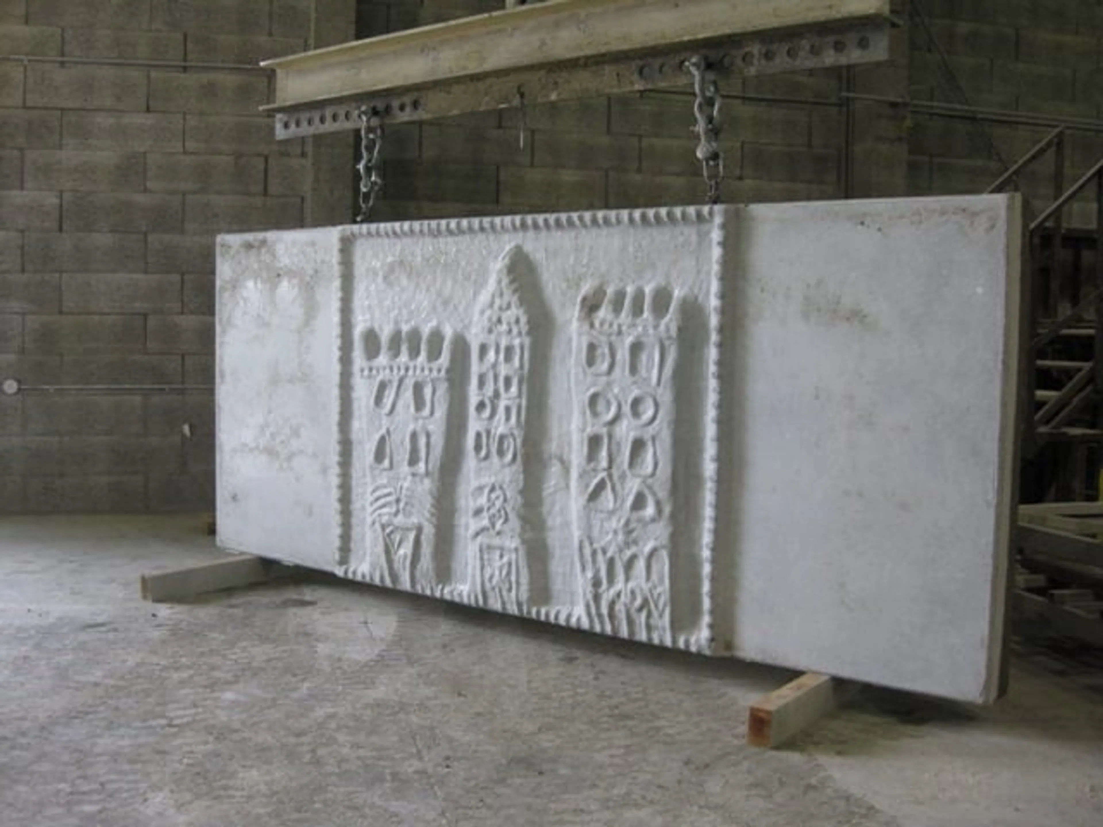 Unpainted precast panel carrying a cityscape relief, suspended from a lifting beam in the plant