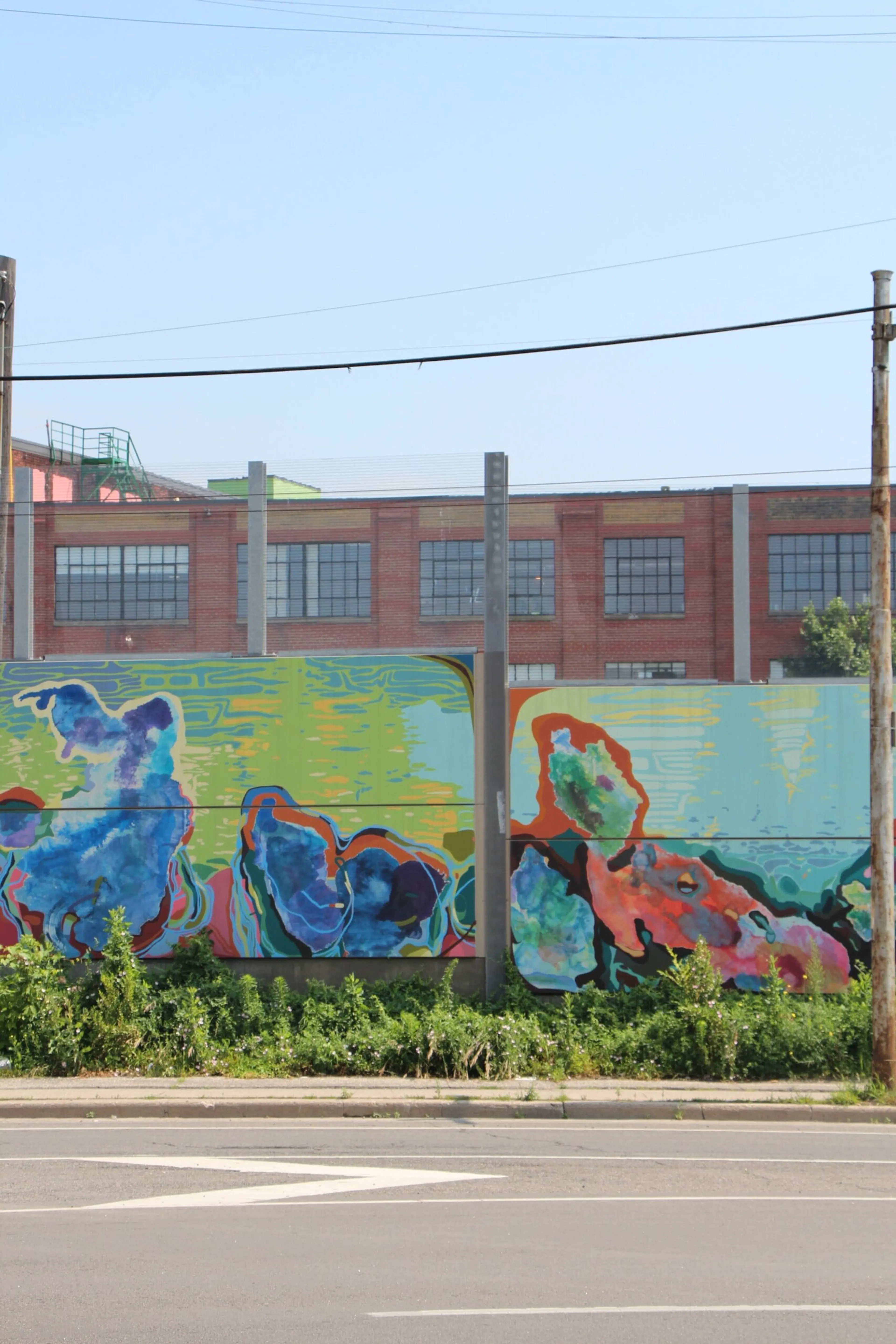 Painted mural panels on the noise wall in front of a brick warehouse along the rail corridor