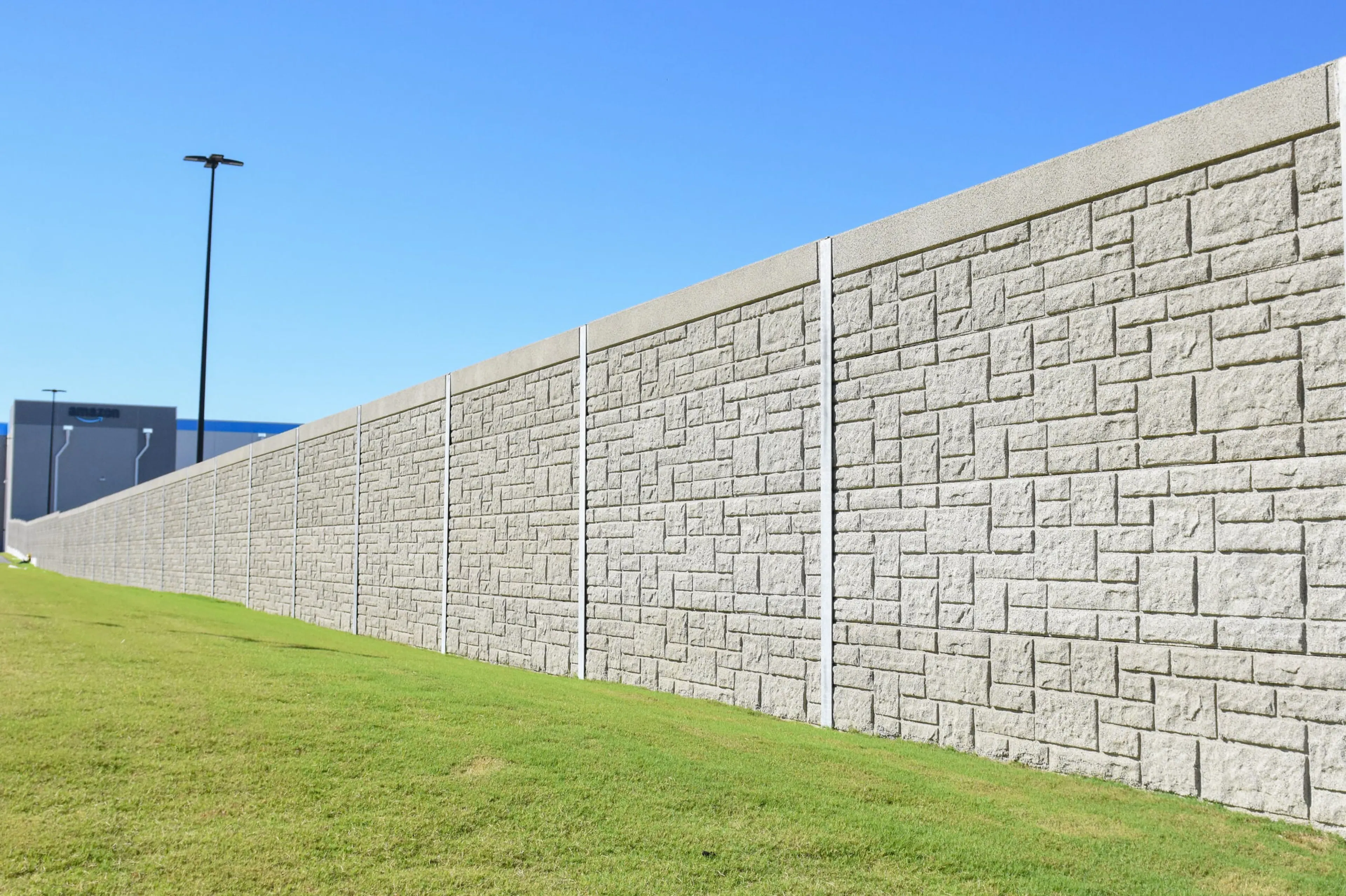 Stone-textured EKHO NB15 noise wall running the length of a lawn toward the Amazon fulfillment center