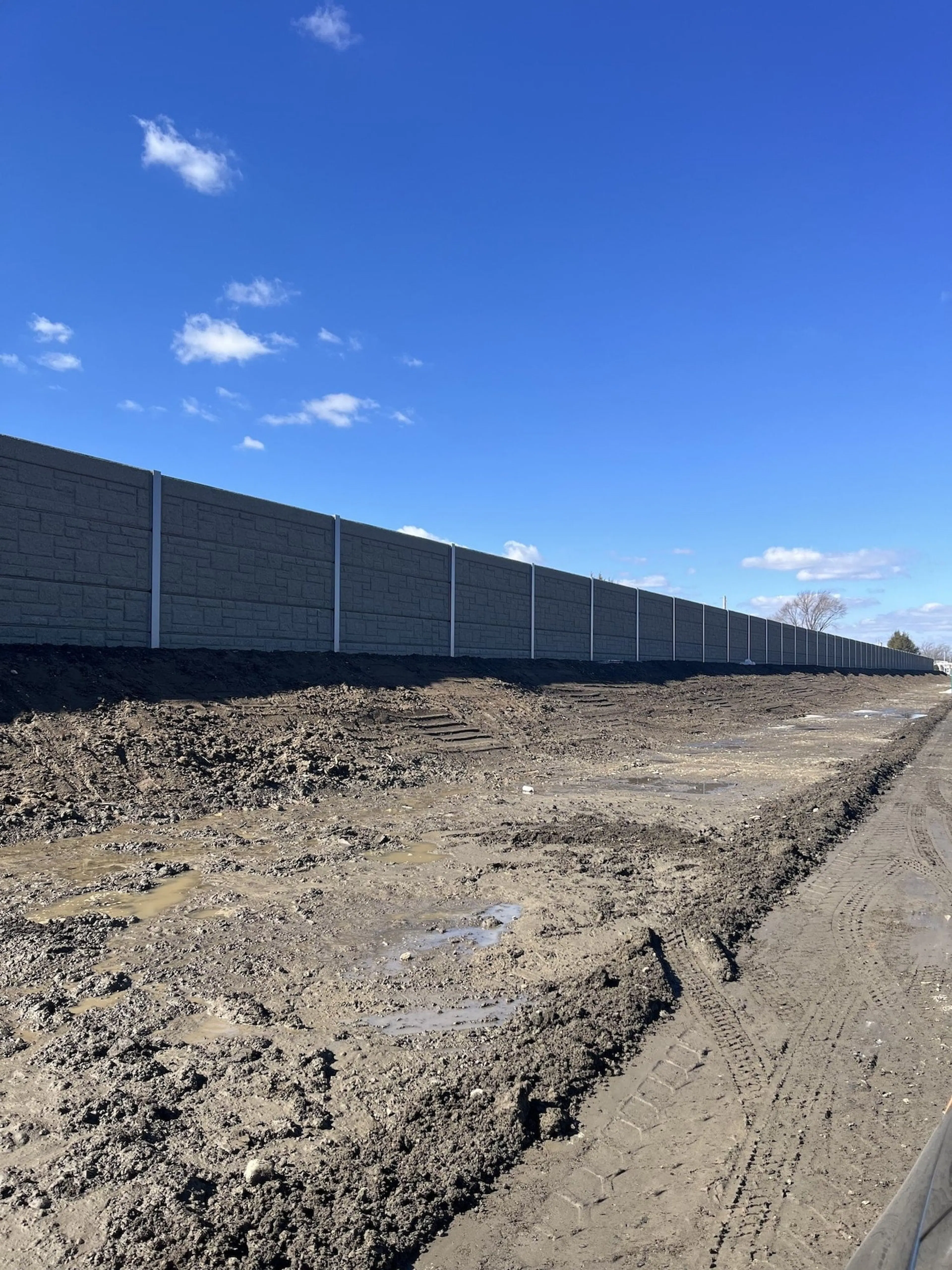 Completed dark grey sound wall behind churned, freshly graded ground at the airport perimeter