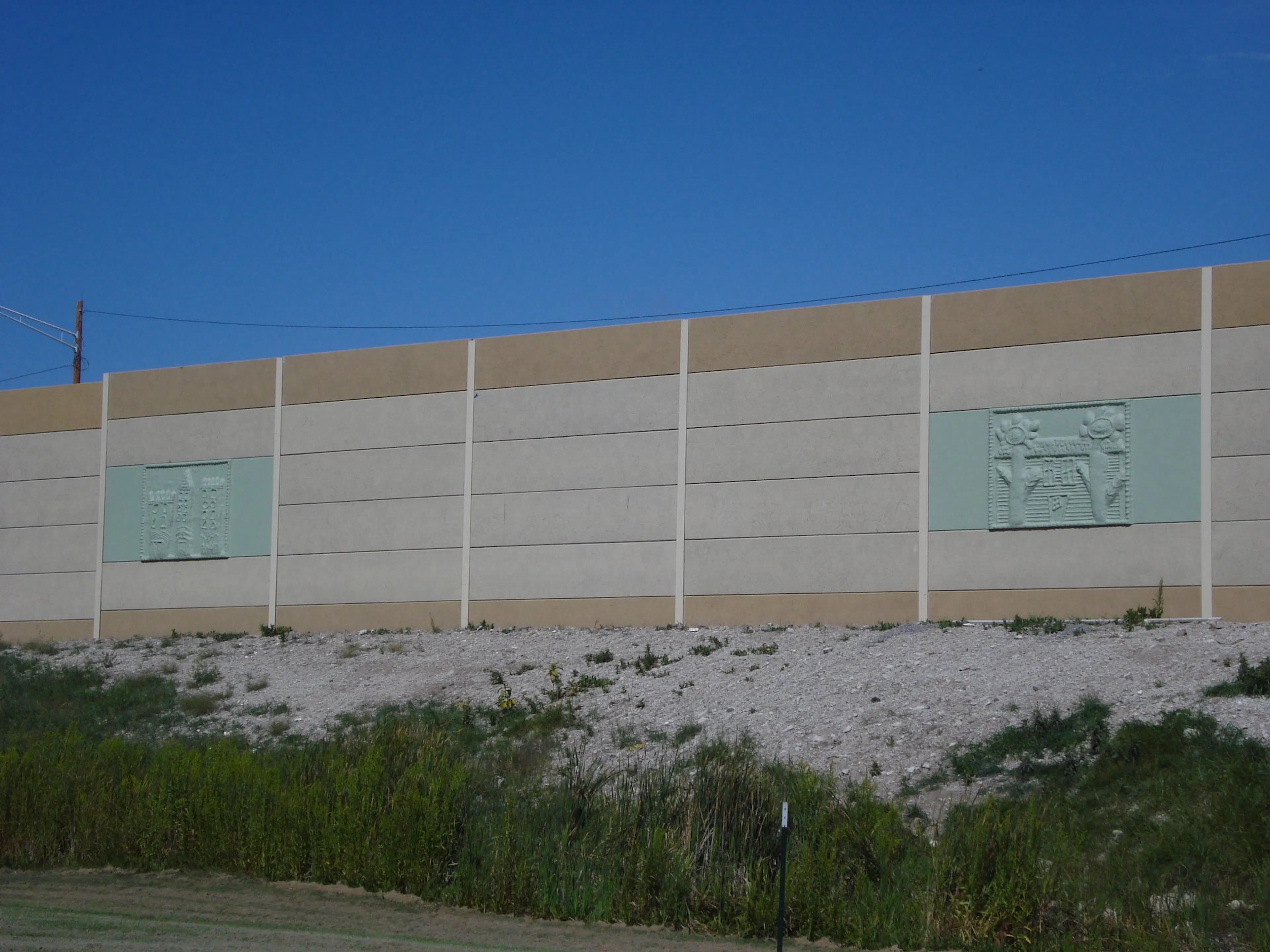 Two pale green icon panels set into a tan noise wall, seen from across the roadside embankment
