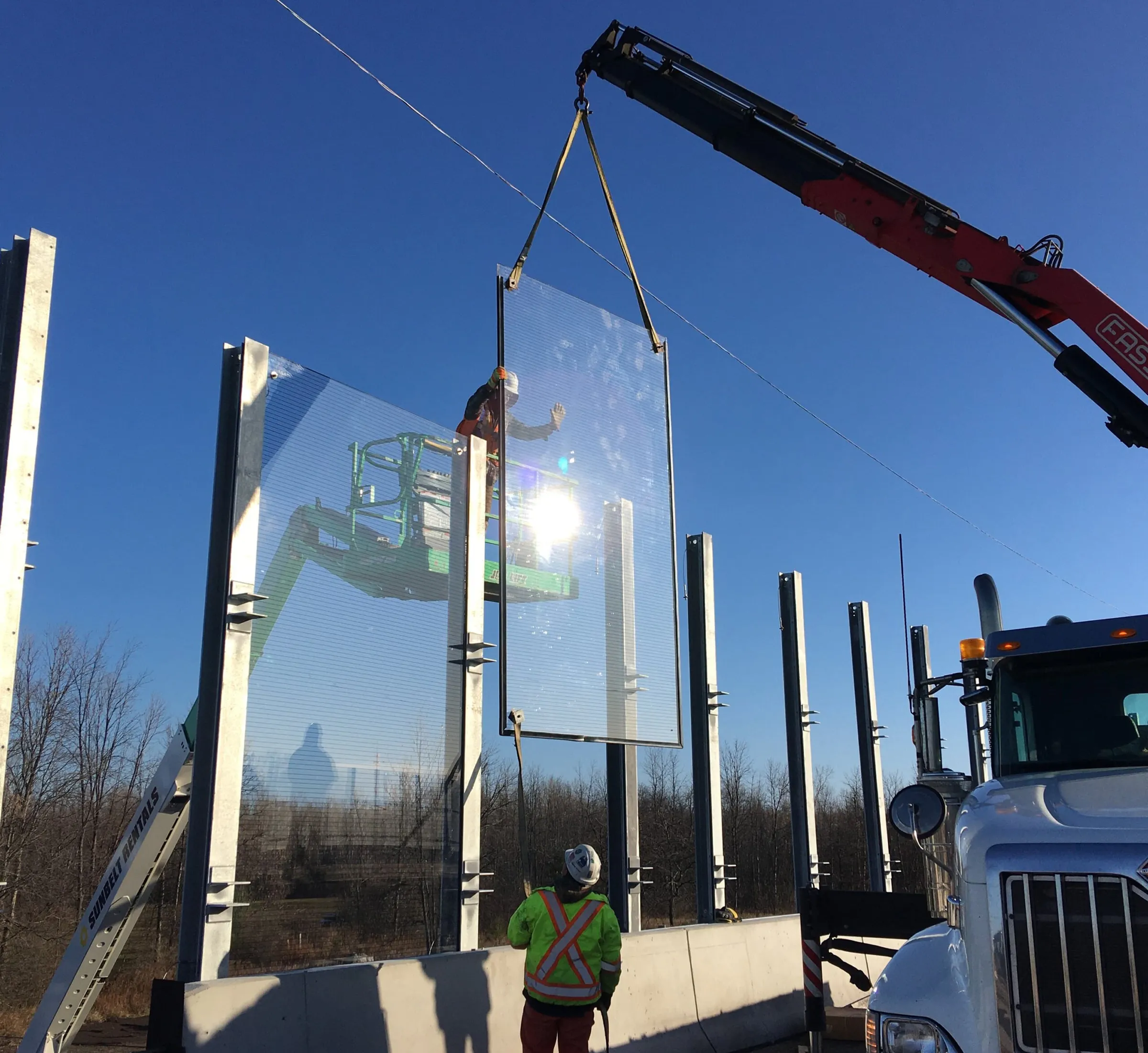 Crane lowering a clear acrylic noise panel between precast posts while a worker in high-visibility gear guides it from below
