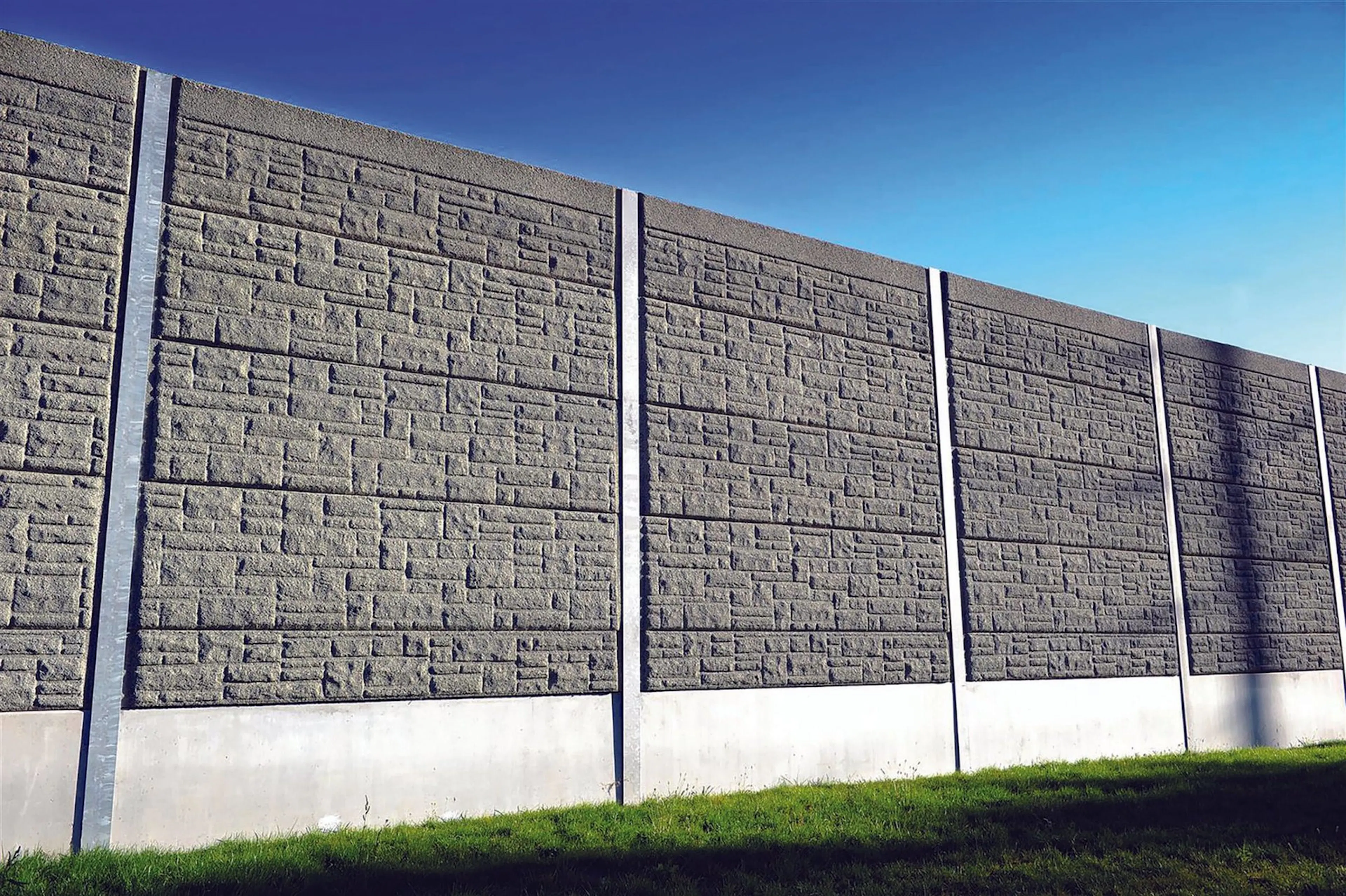 Precast absorptive panels stacked between posts, the barrier rising above a grass slope against a blue sky