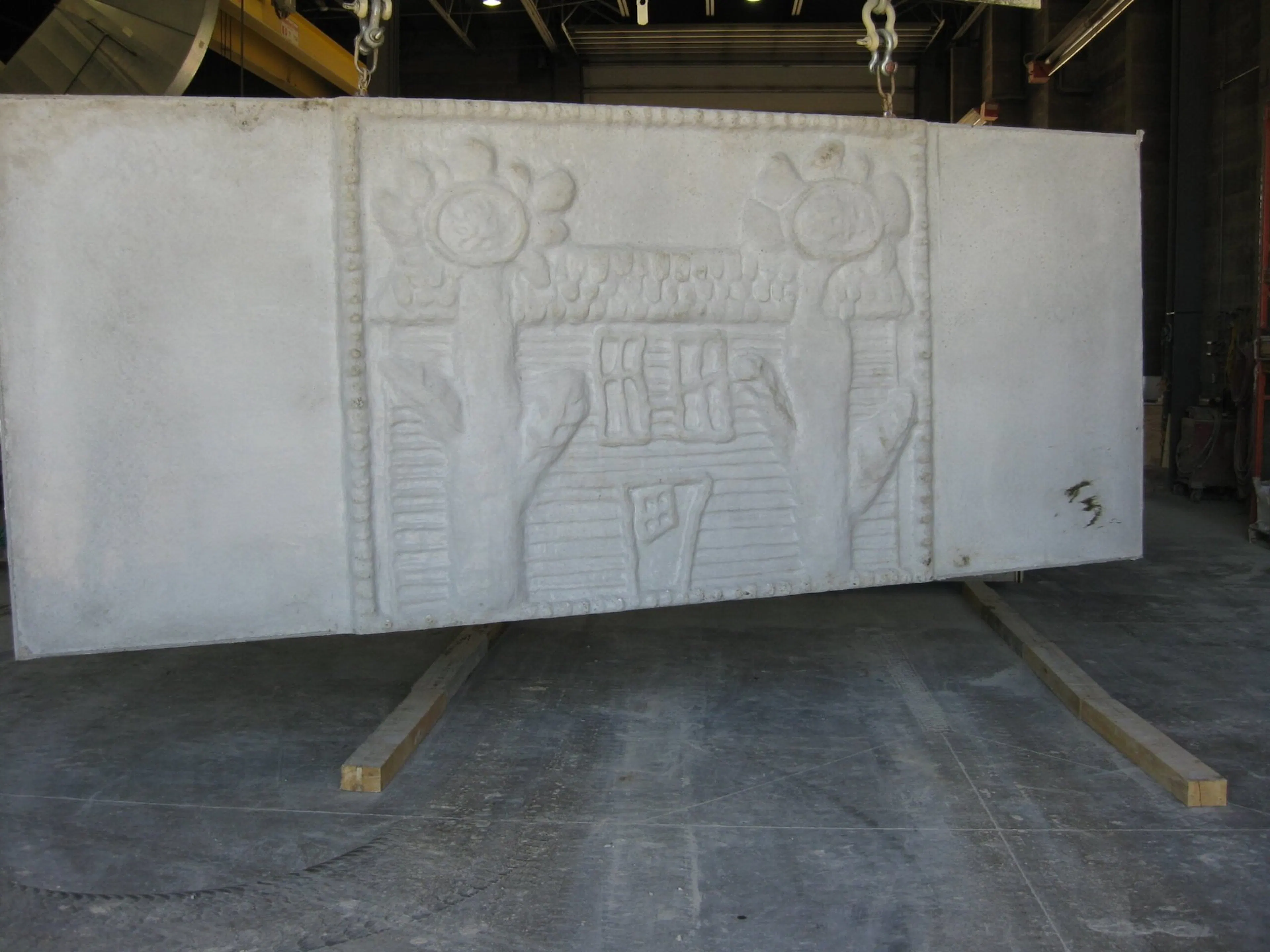 Unpainted precast panel carrying the house and sunflowers relief, lifted by chains inside the plant