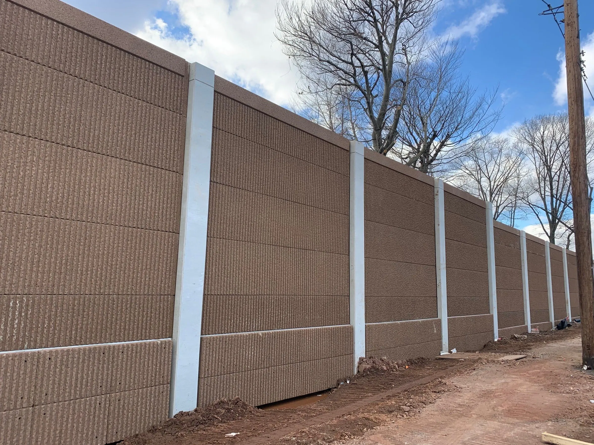 Brown absorptive noise wall with a vertical ribbed finish set between white posts along a cleared roadside