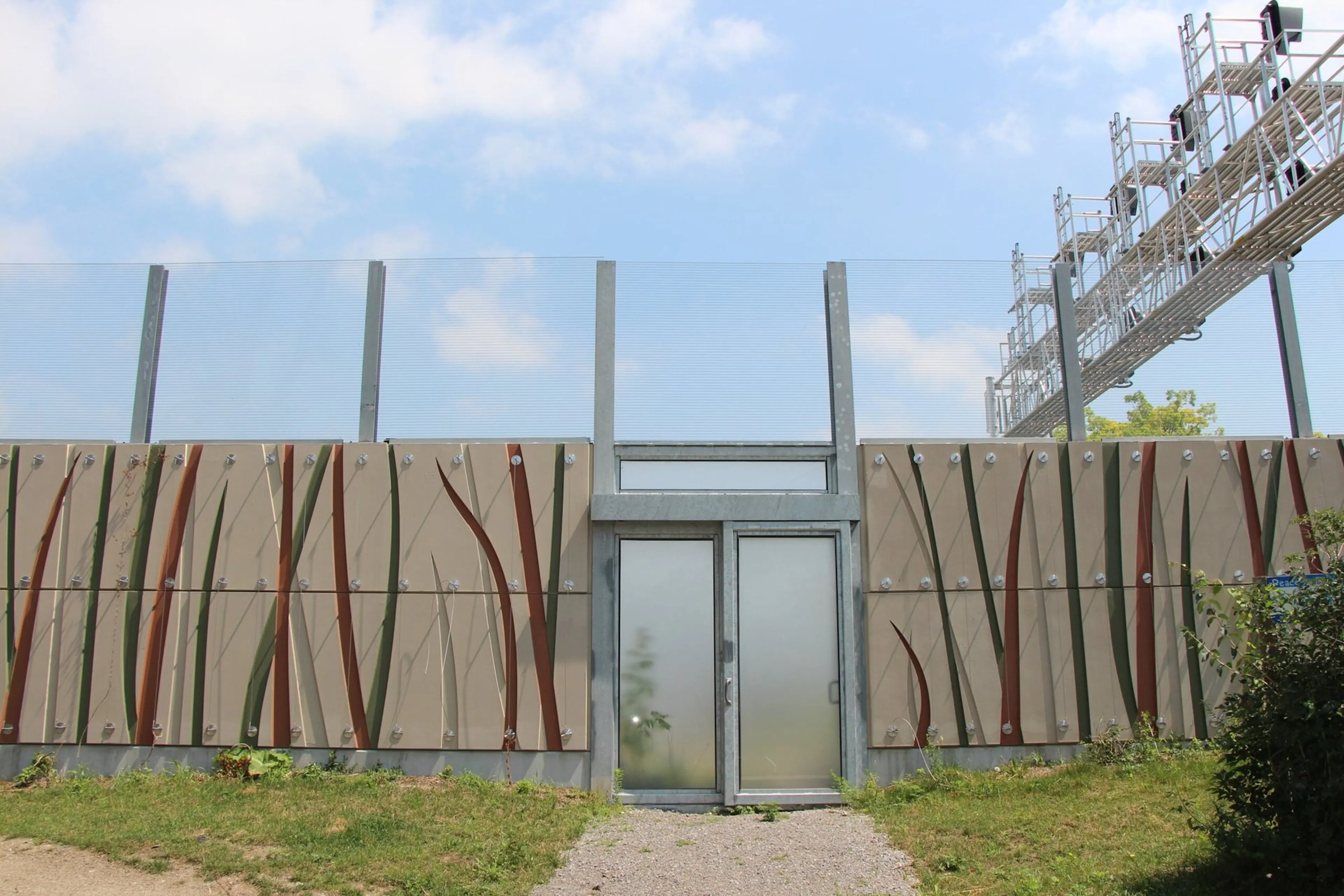 Noise wall with cast reed patterns and a glazed access gate, a rail signal gantry rising behind