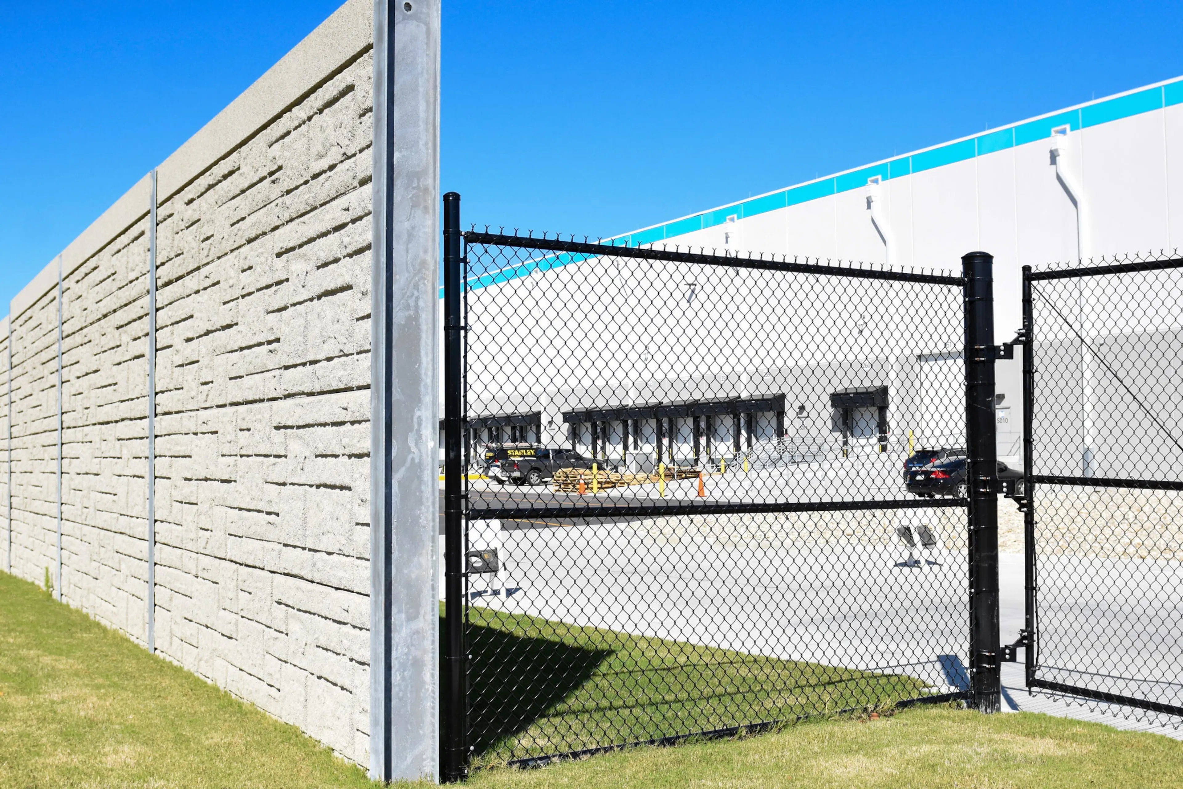 Ribbed rear face of the noise wall meeting a black chain-link gate, with the fulfillment center loading docks beyond