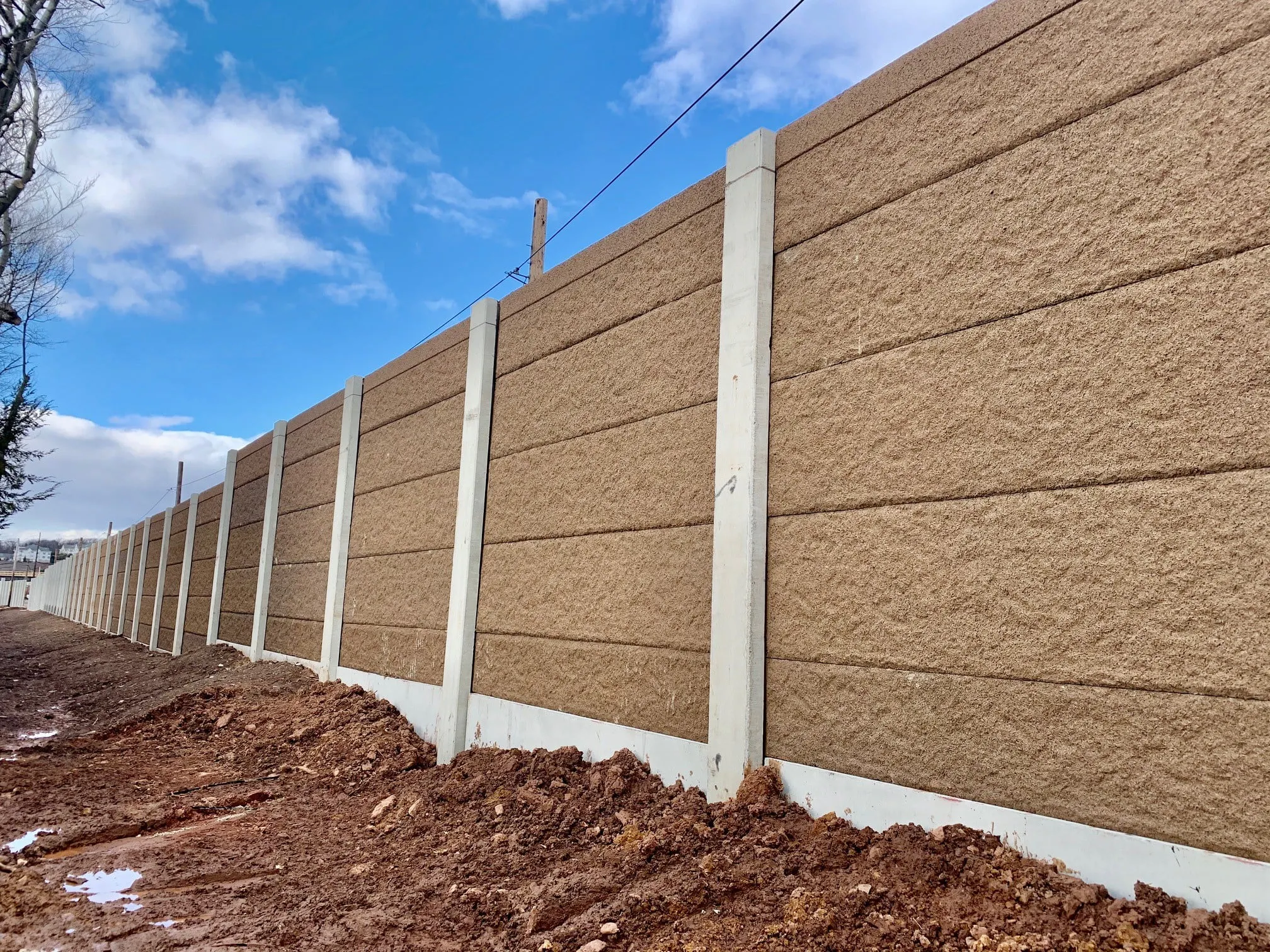 Sand-toned absorptive noise wall panels stacked between concrete posts above a freshly graded embankment