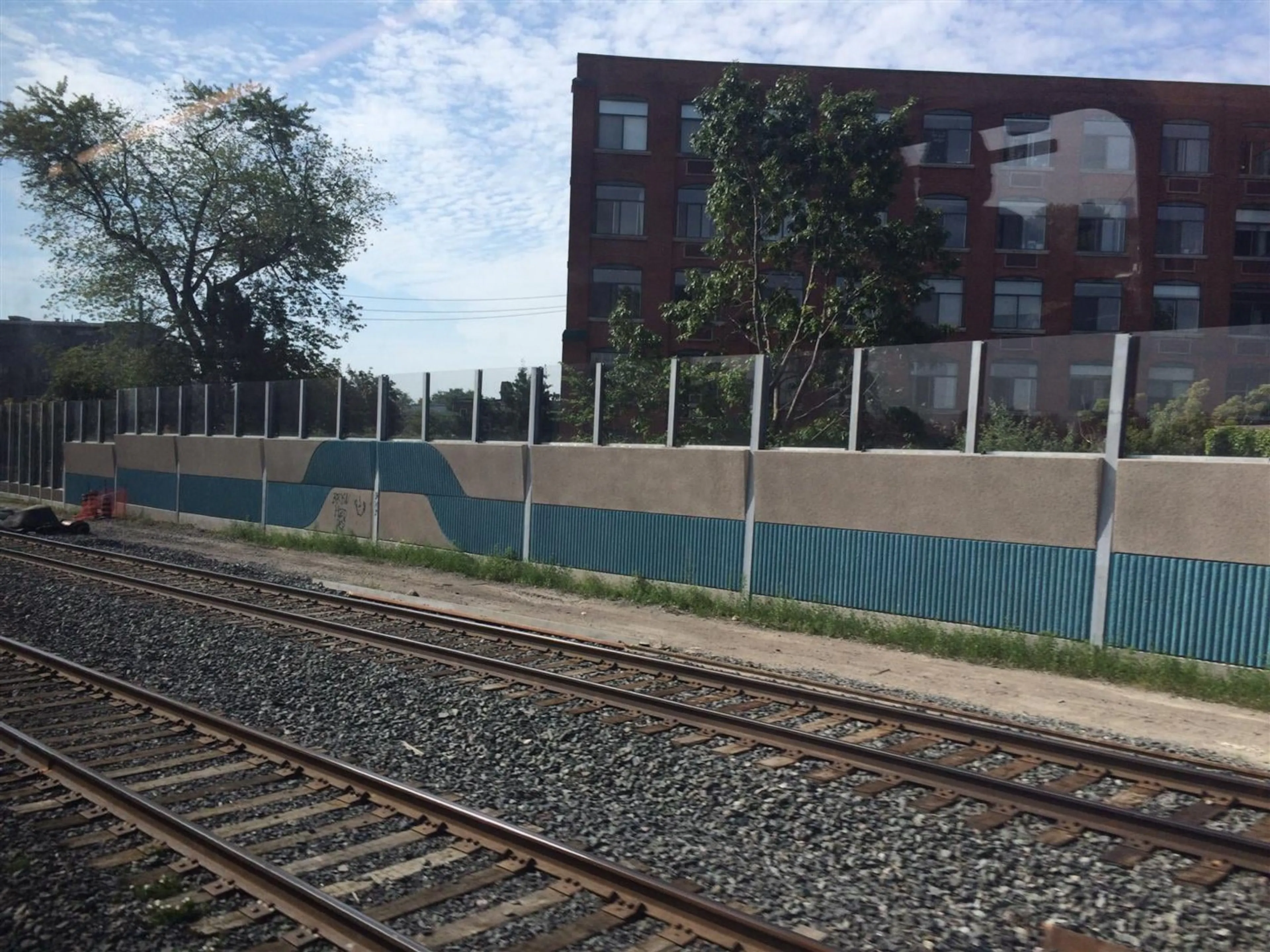 Teal wave pattern along the UP Express wall, seen across the tracks with a brick industrial building behind