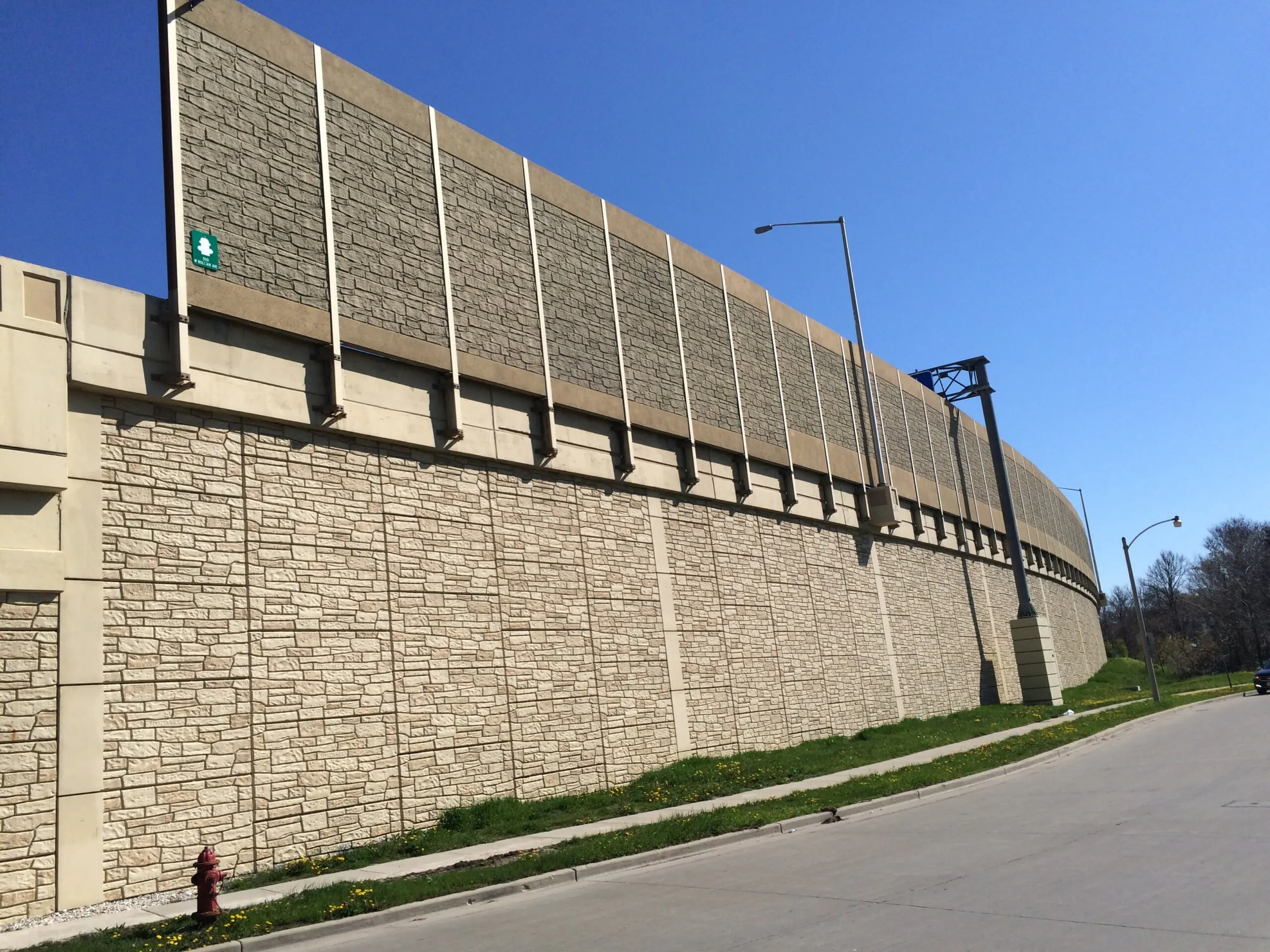 Tall stone-textured noise wall curving along a Milwaukee roadway above a matching retaining wall