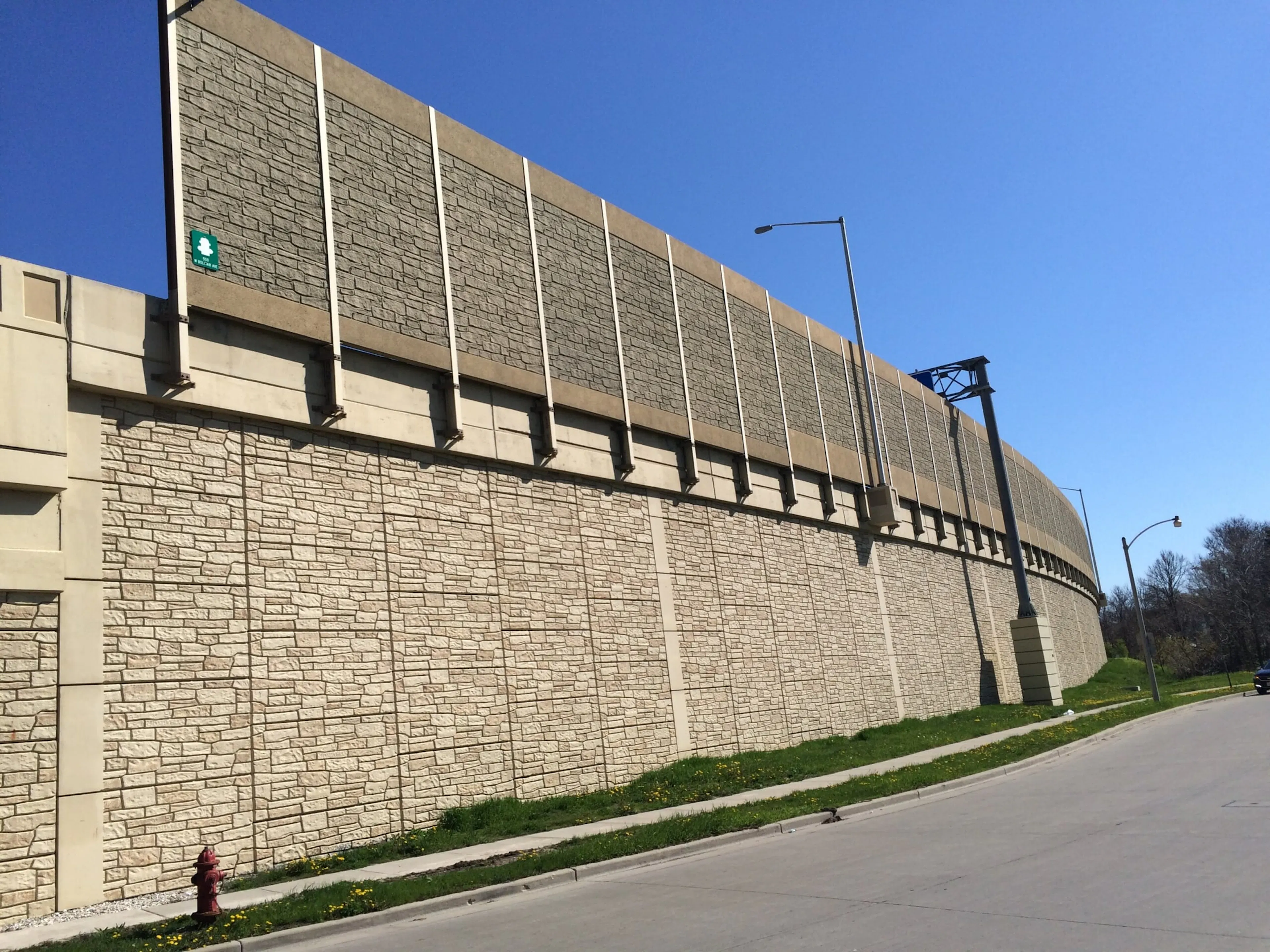 Tall stone-textured noise wall curving along a Milwaukee roadway above a matching retaining wall