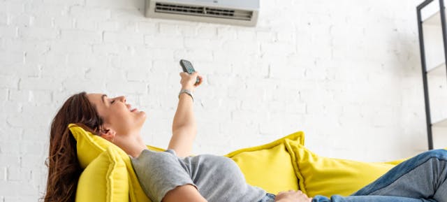 Une femme allongée sur son canapé jaune allume la climatisation de sa maison à l'aide d'une télécommande