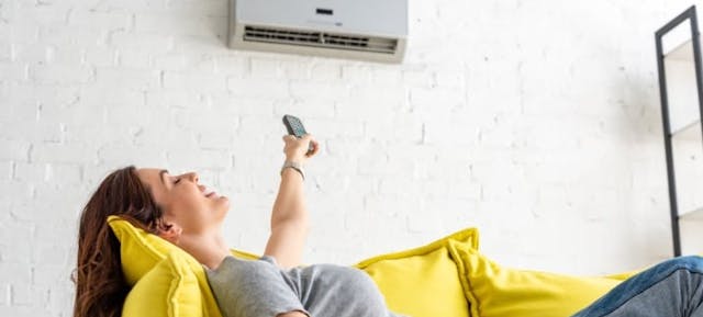 Une femme allongée sur son canapé jaune allume la climatisation de sa maison à l'aide d'une télécommande