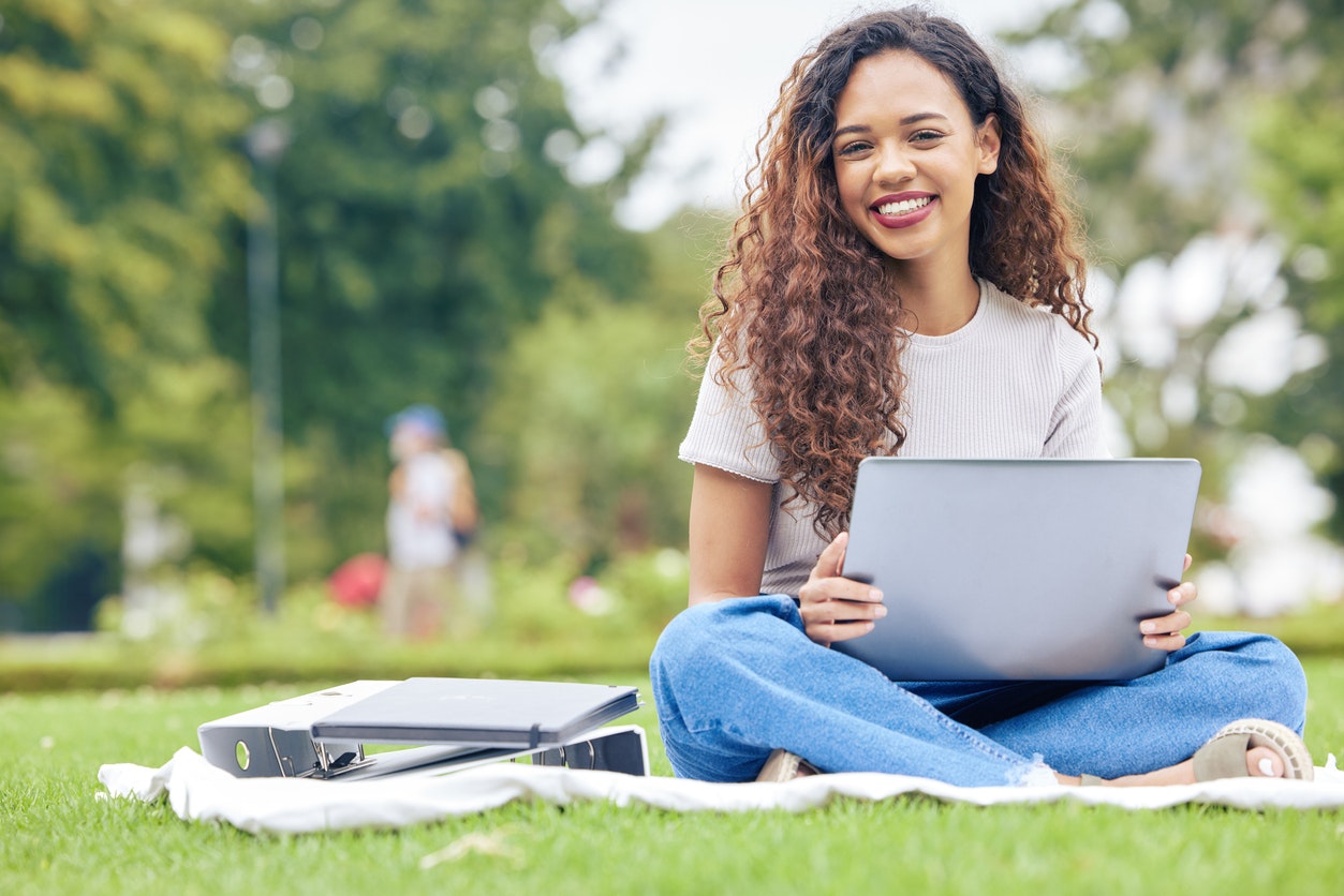 Changing Student Expectations. One young woman working on her laptop while sitting outside on an open field. A beautiful female student smiling while using her computer to study online on her university campus stock photo