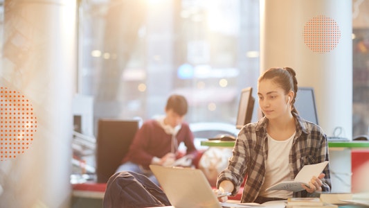 Young white female with brown hair sitting at a laptop computer with a notebook in hand.