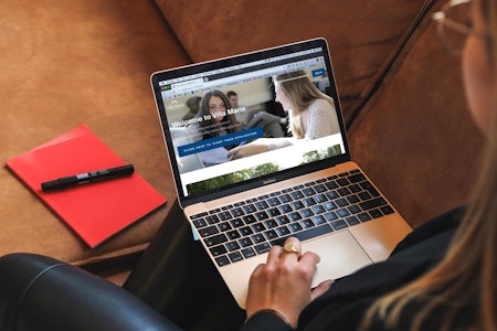Picture of a woman browsing the Villa Maria website on her laptop, and having a red notebook next to her on the sofa.